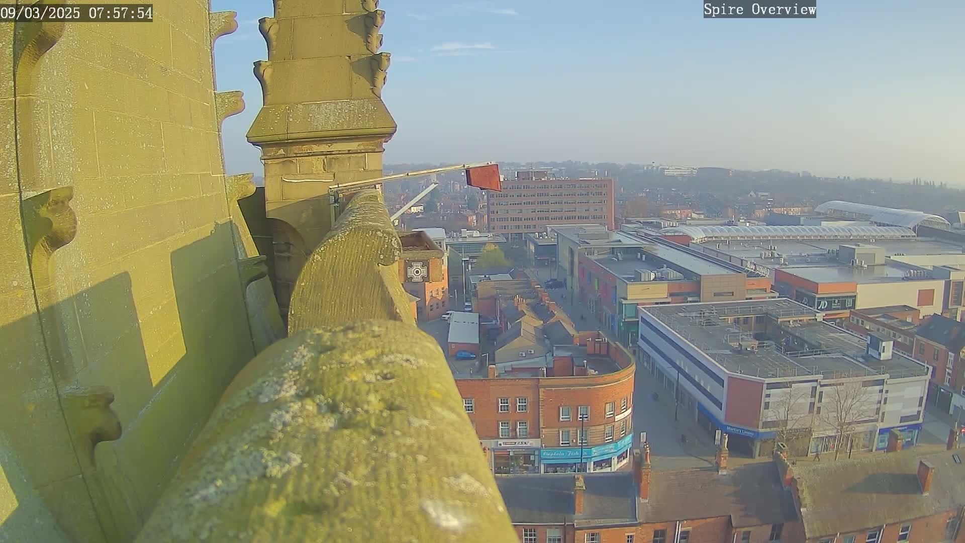 A high-angle, sunny view from a stone structure shows a town square with buildings and a shopping center below.