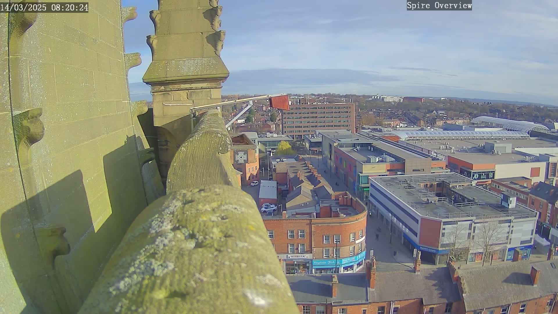 A high-angle view from a stone structure on a sunny day shows a town square with shops and buildings, including a large shopping center with a glass roof.