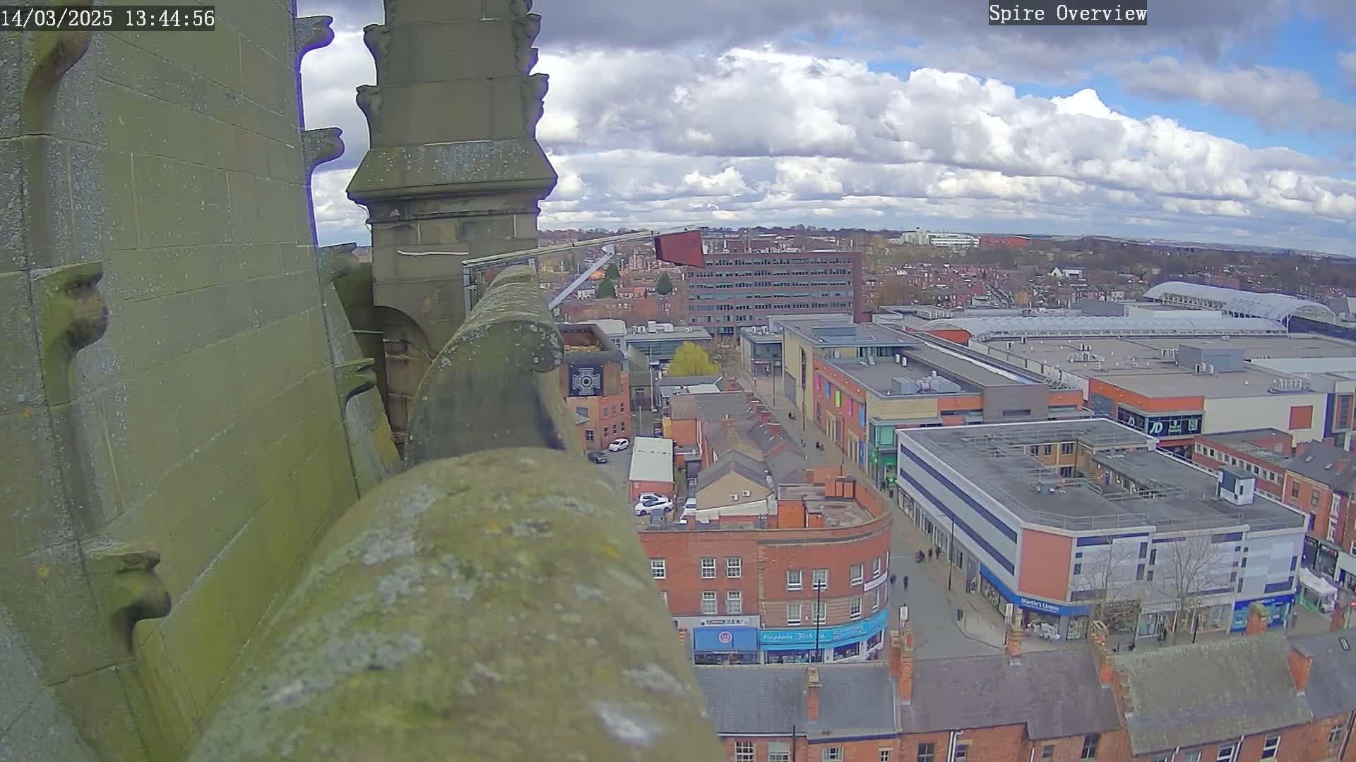 A high-angle view from a stone structure shows a town with partly cloudy skies, overlooking a shopping center and residential buildings.