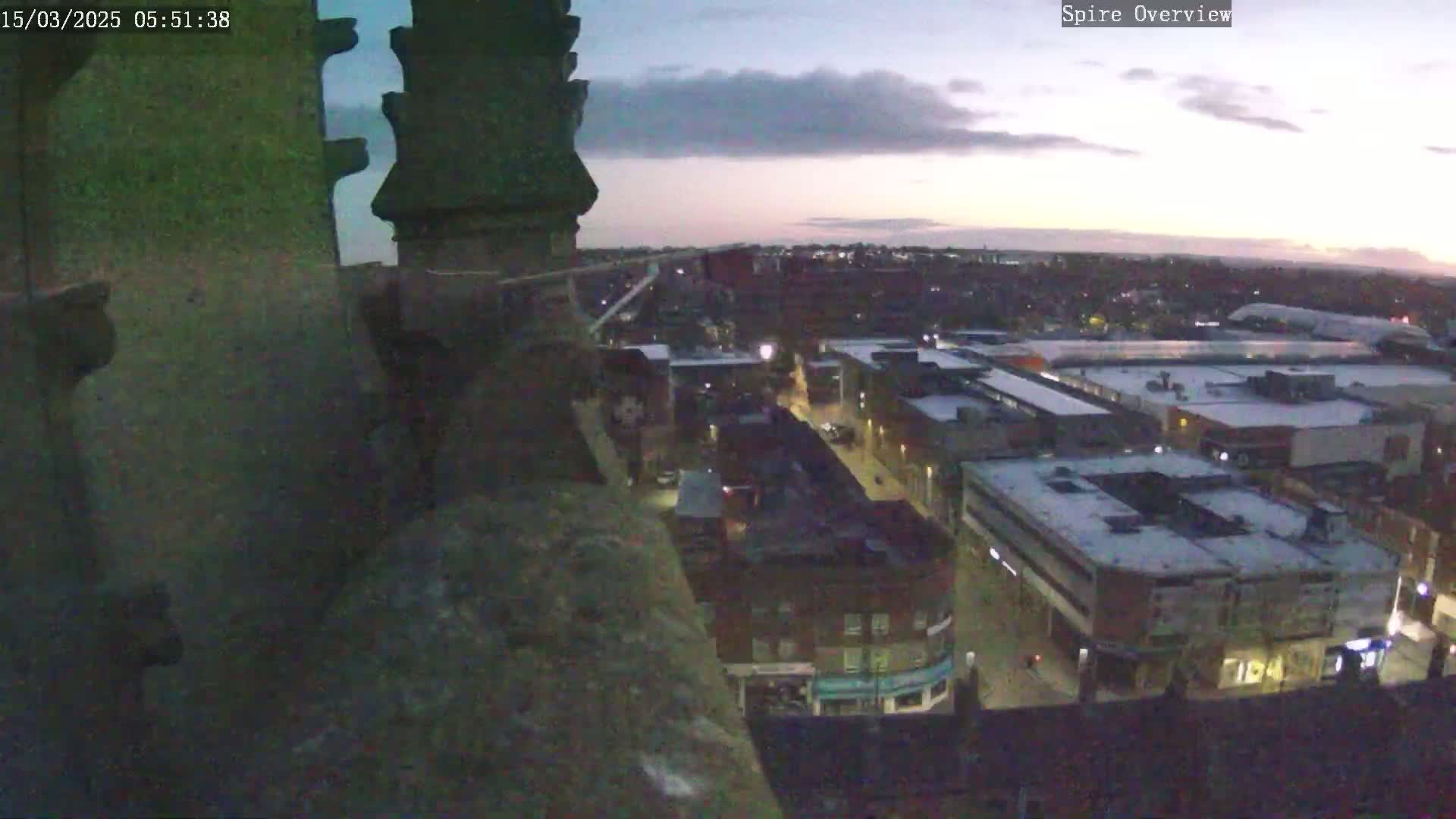 A twilight view from a high vantage point shows a town with low-lying buildings and streets, some of which are covered in a light dusting of snow.