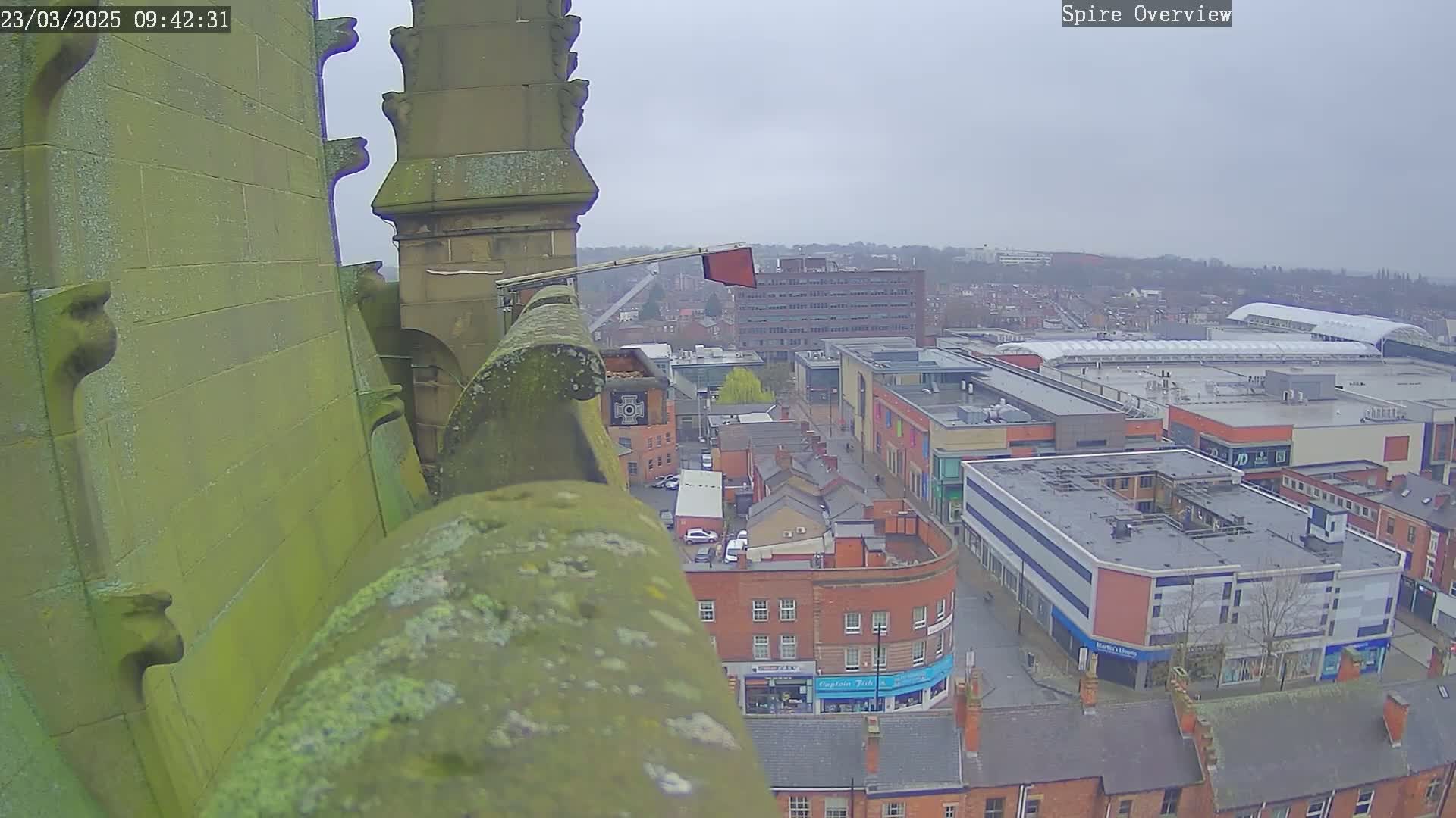 A high-angle view from a stone structure shows an overcast day over a town square with brick buildings and a large shopping center.
