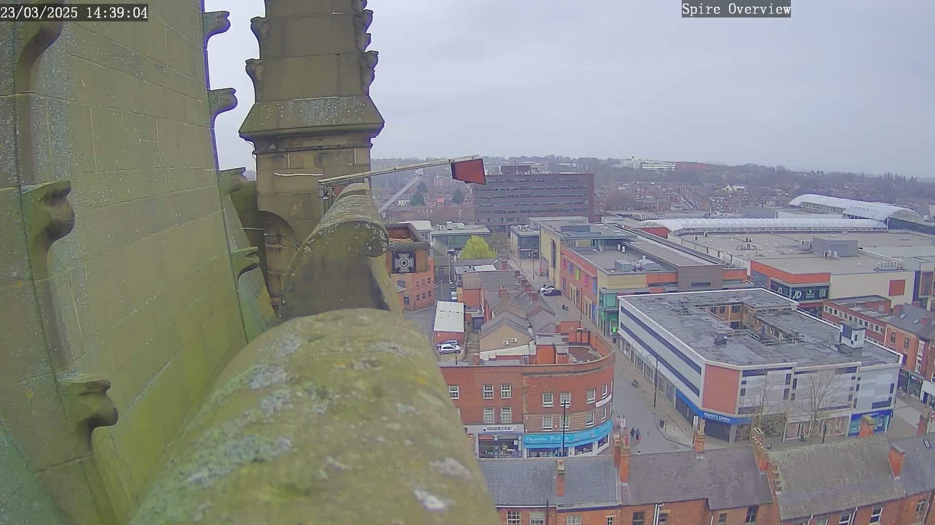 A high-angle view from a stone spire shows an overcast day over a town with a large shopping center.