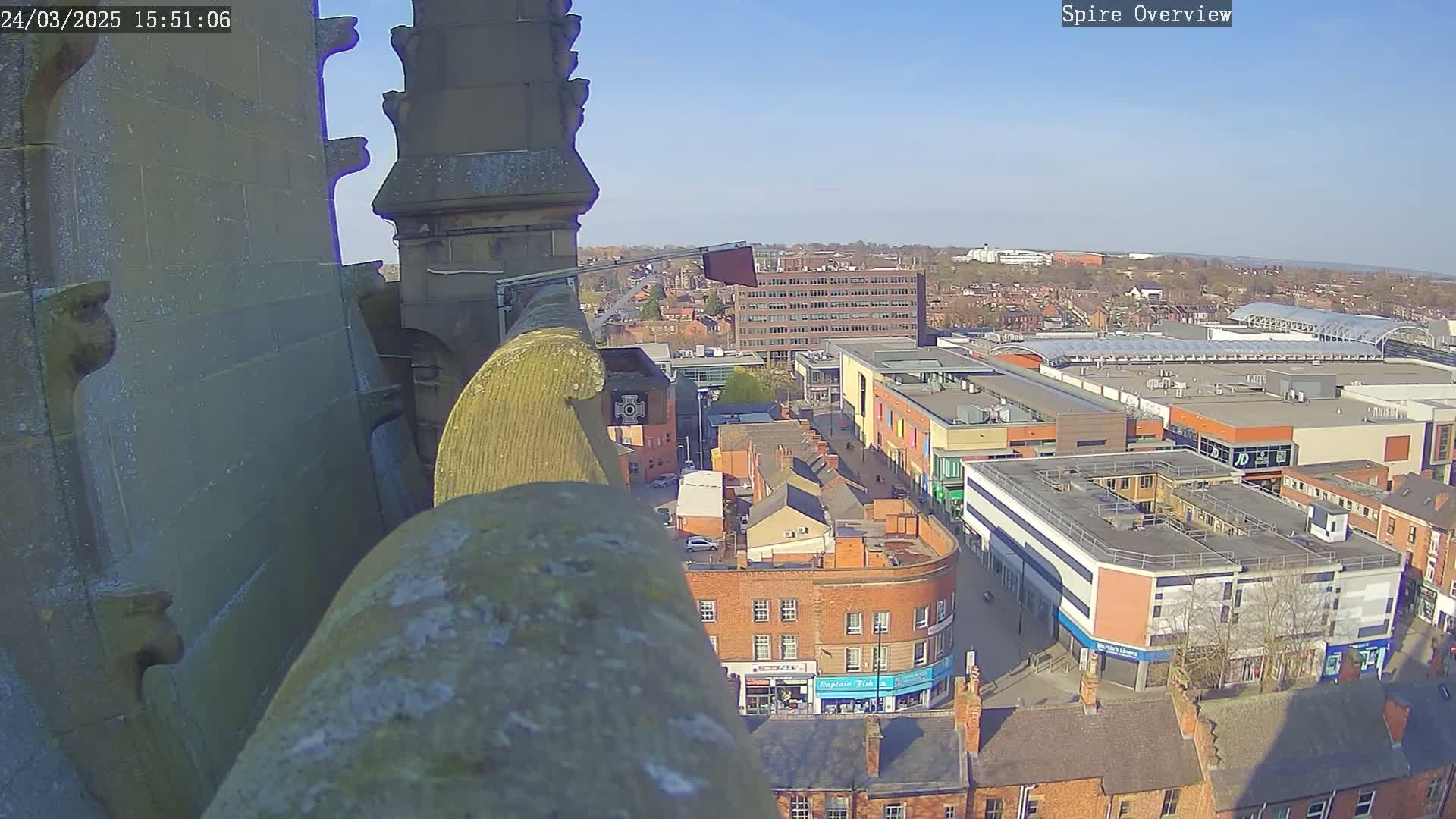 A high-angle view from a stone structure shows a town square on a sunny day, with buildings and a shopping center visible in the distance.