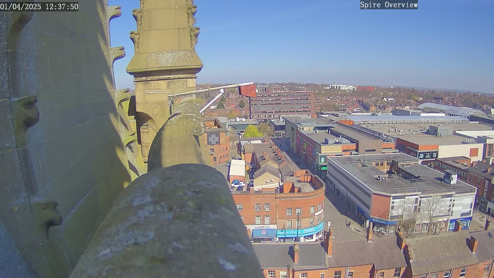 A high-angle, sunny view from a stone spire shows a town square with a shopping center and surrounding buildings.