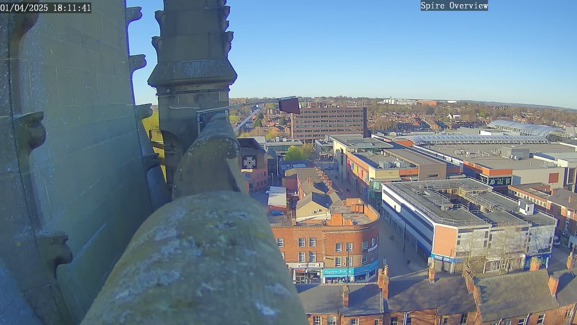 A high-angle view from a stone structure shows a town square with buildings and a shopping center under a clear, sunny sky.