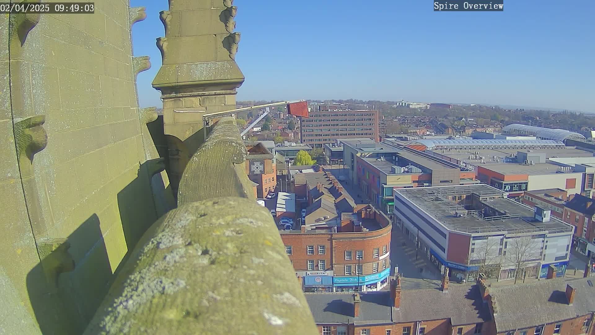 A high-angle view on a sunny day shows a stone structure overlooking a town square with shops and buildings, including a large multi-story building.