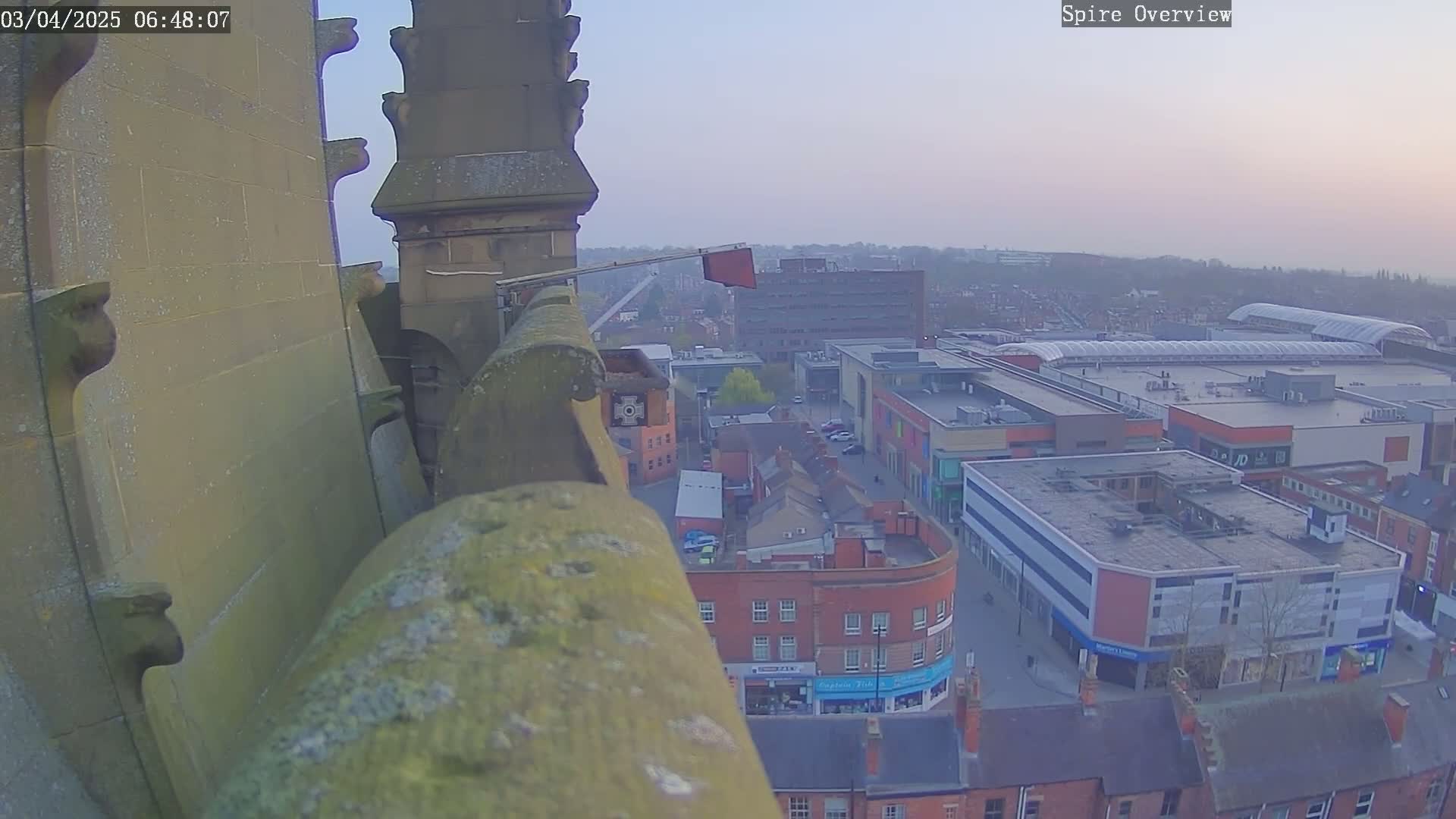 A hazy sunrise view from a high vantage point shows a stone structure overlooking a town with a large shopping center and numerous buildings.