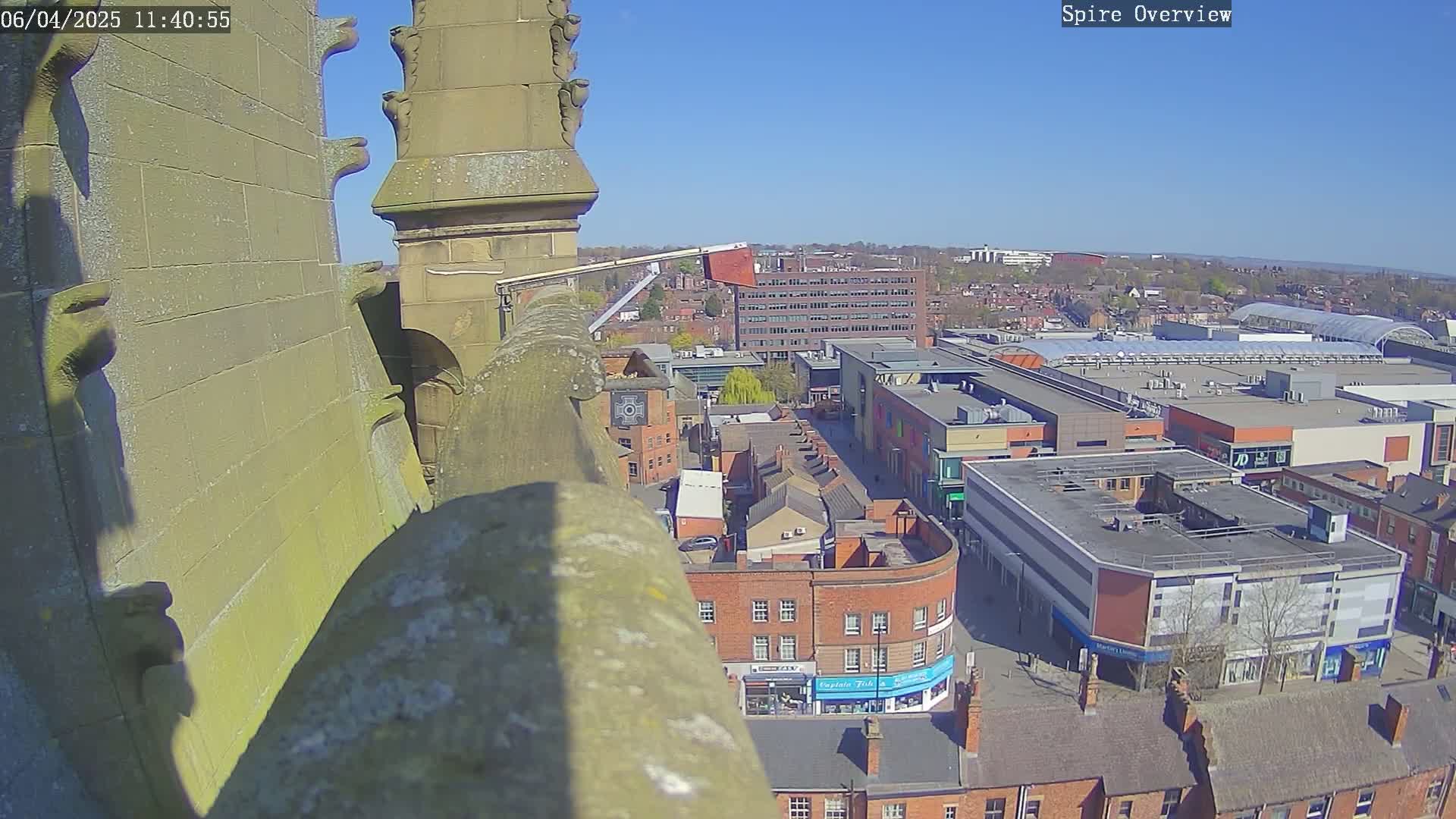 A high-angle view on a clear, sunny day shows a stone structure overlooking a town square with shops and buildings.