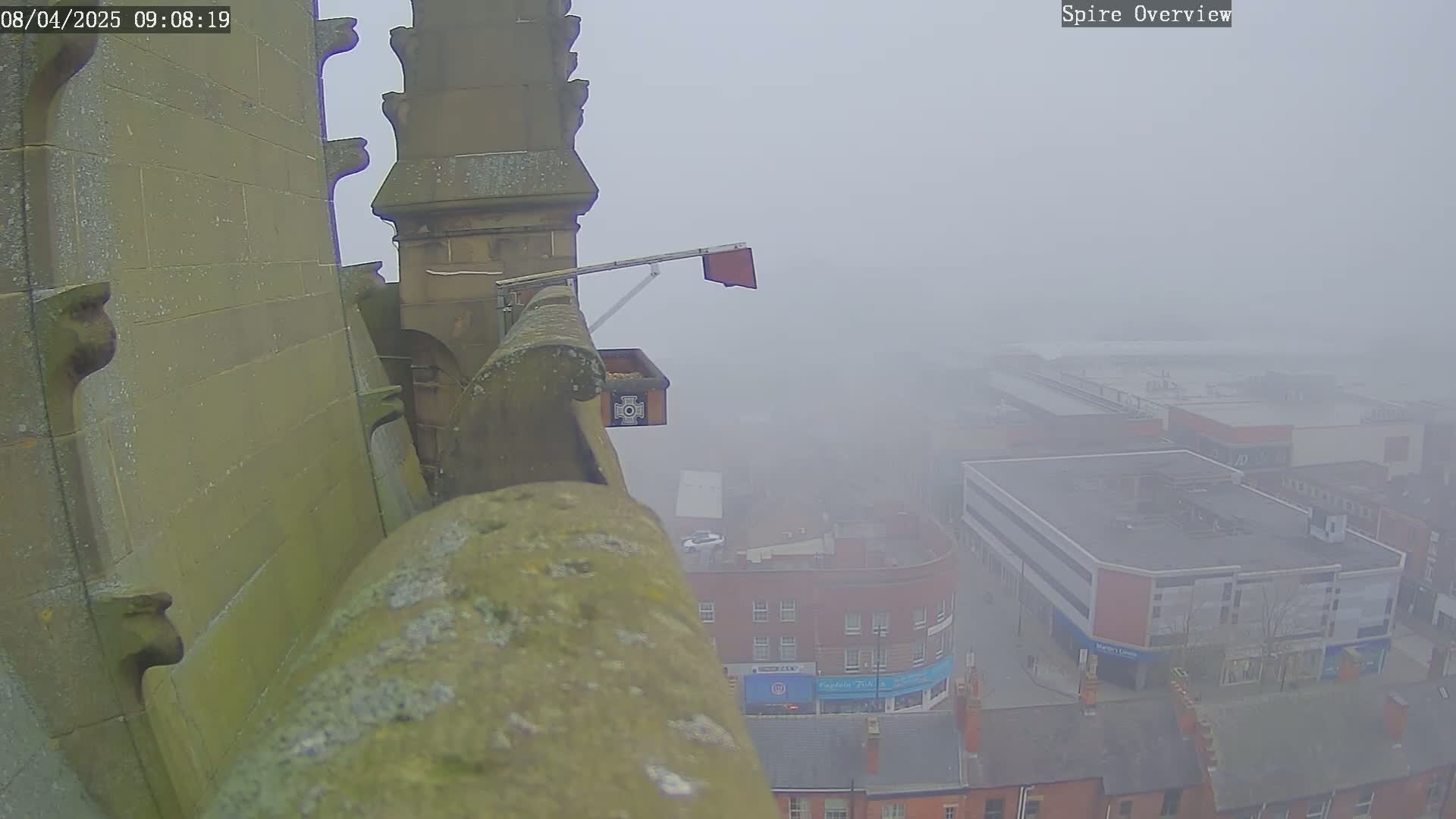 A foggy view from a high vantage point shows a stone structure with a bird feeder, overlooking a town square with buildings obscured by mist.