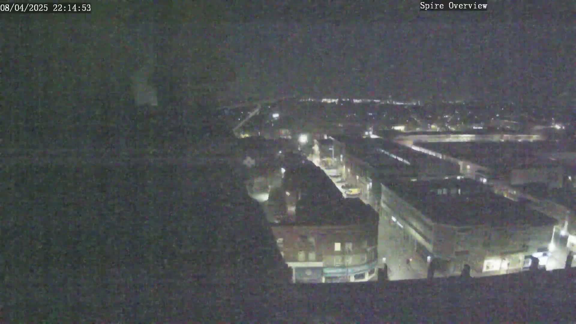 A nighttime, high-angle view shows a city block with several buildings and a street, extending into a larger area of city lights under a dark sky.