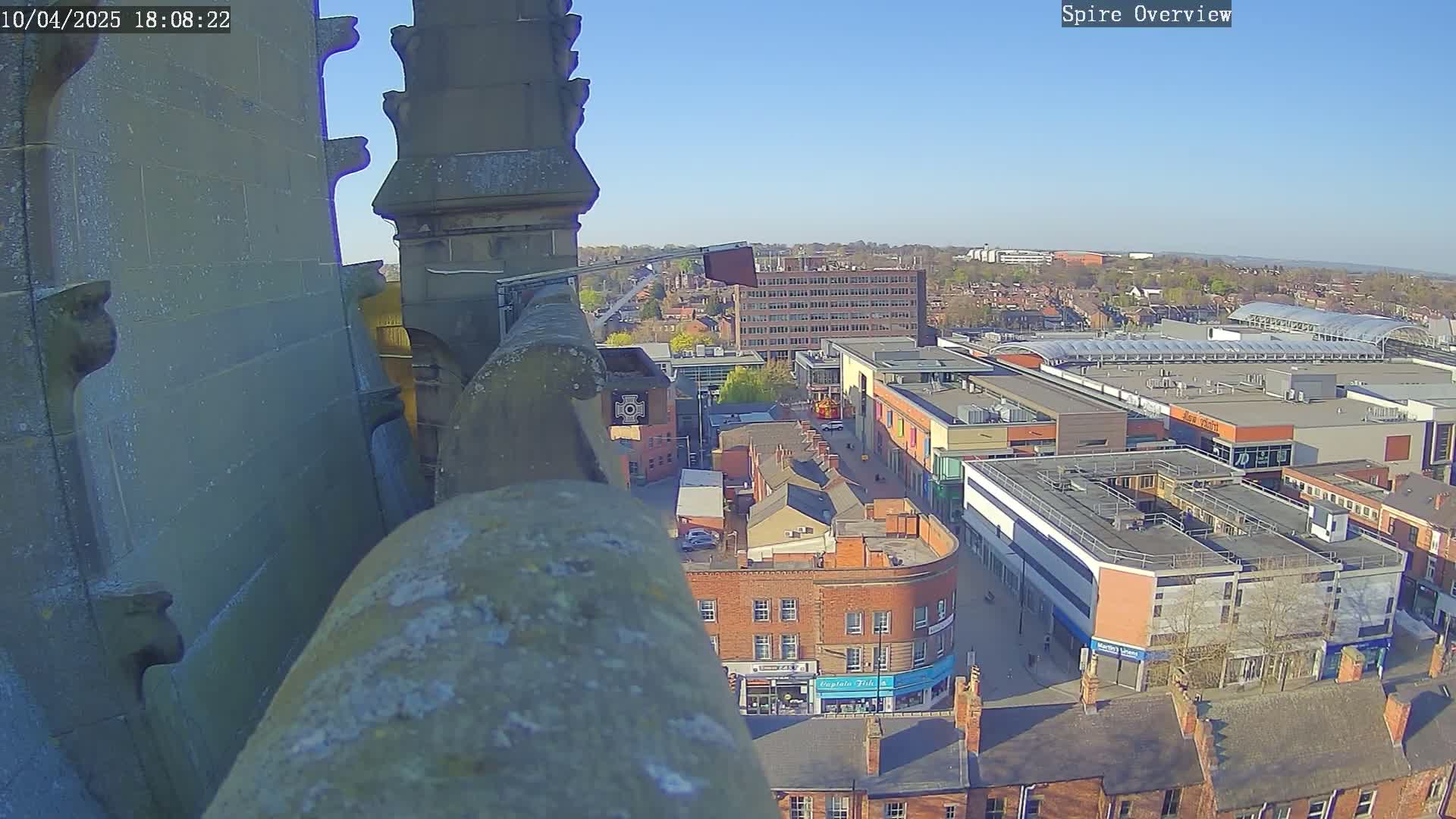 A high-angle view from a stone structure shows a town square with buildings and a shopping center under a clear, sunny sky.