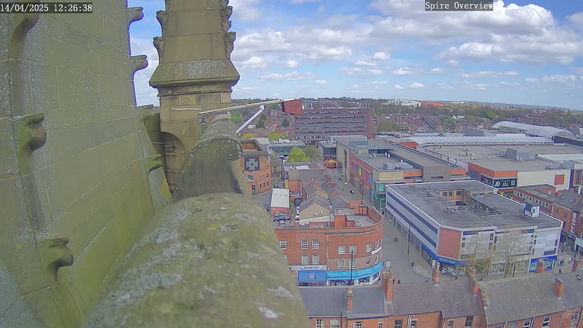 A high-angle view from a stone structure shows a partly sunny day over a town square with brick buildings and a large shopping center, a partly cloudy sky is visible.
