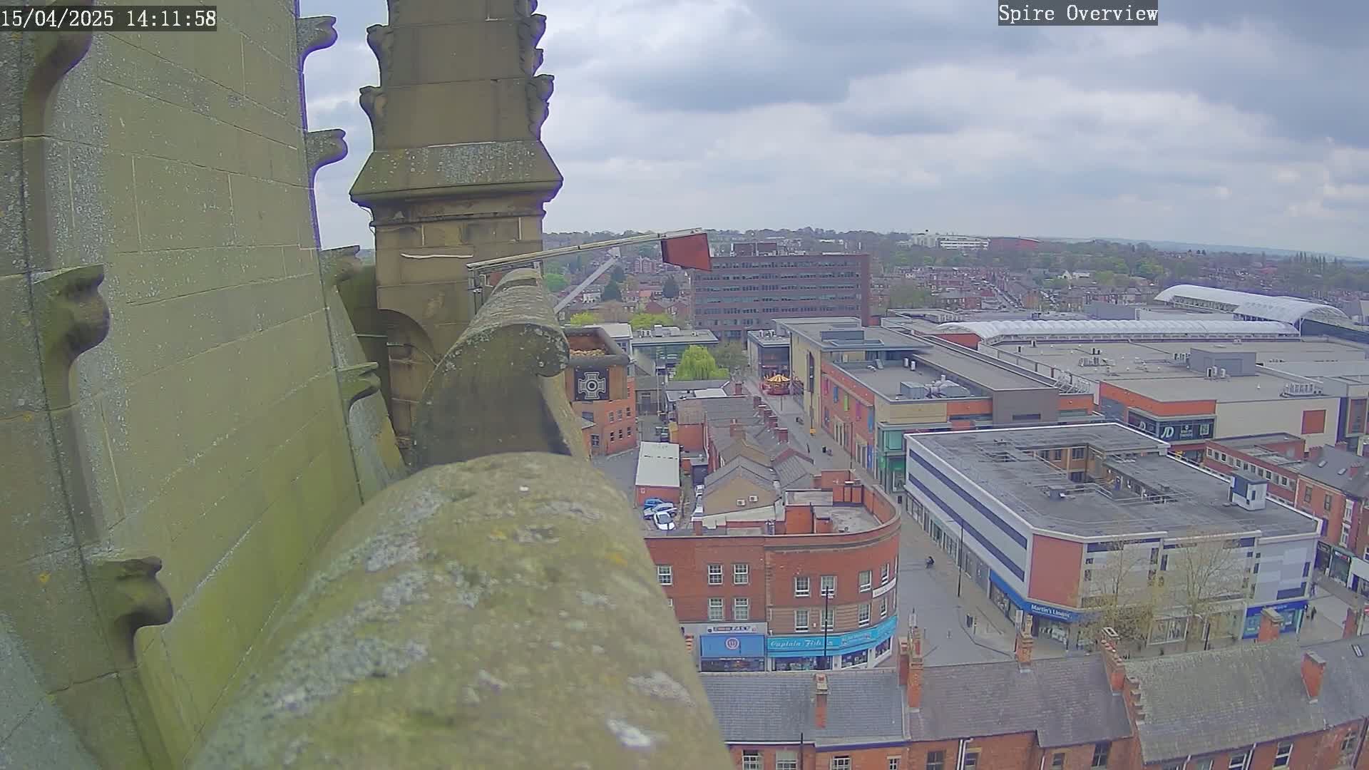 A high-angle view from a stone spire shows an overcast day over a town square with brick buildings and a large shopping center.