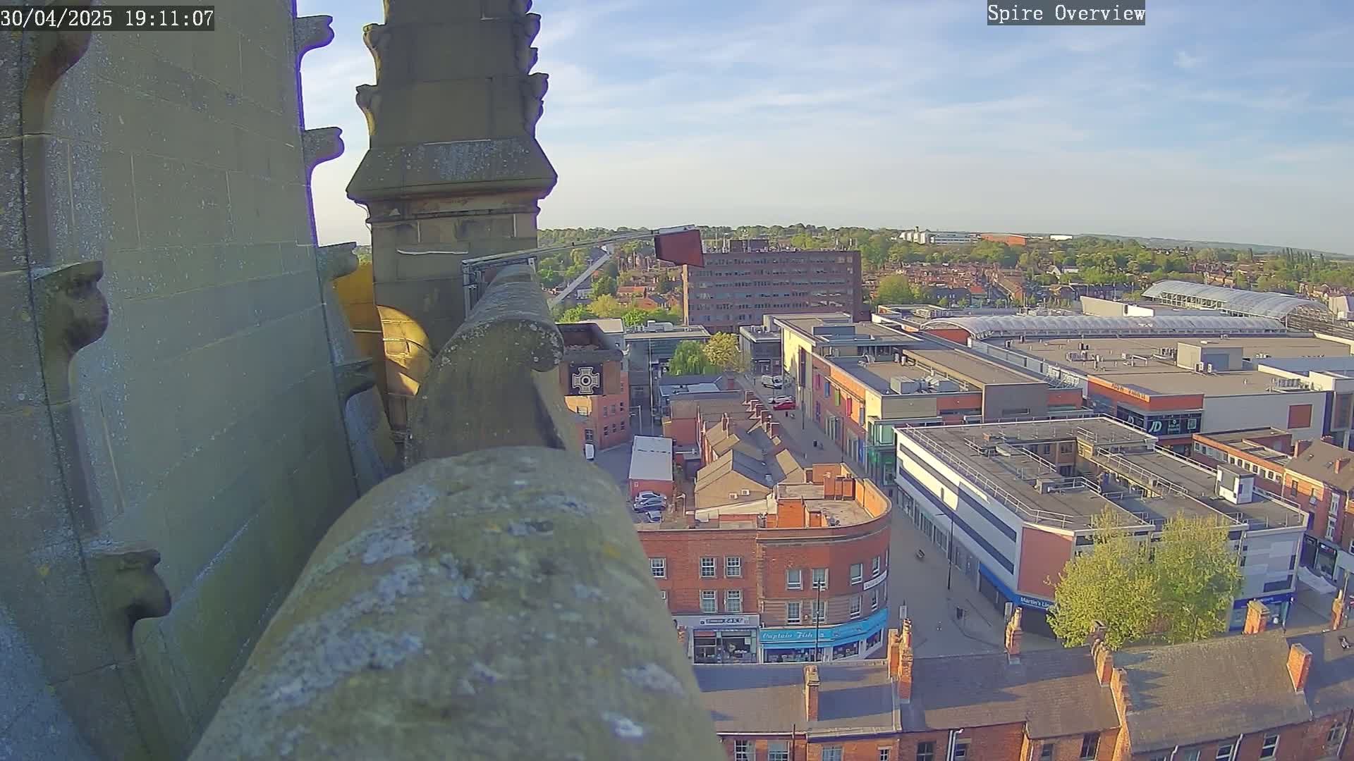 A high-angle view from a stone structure shows a town under a mostly clear, sunny sky, with a large shopping center prominently featured.
