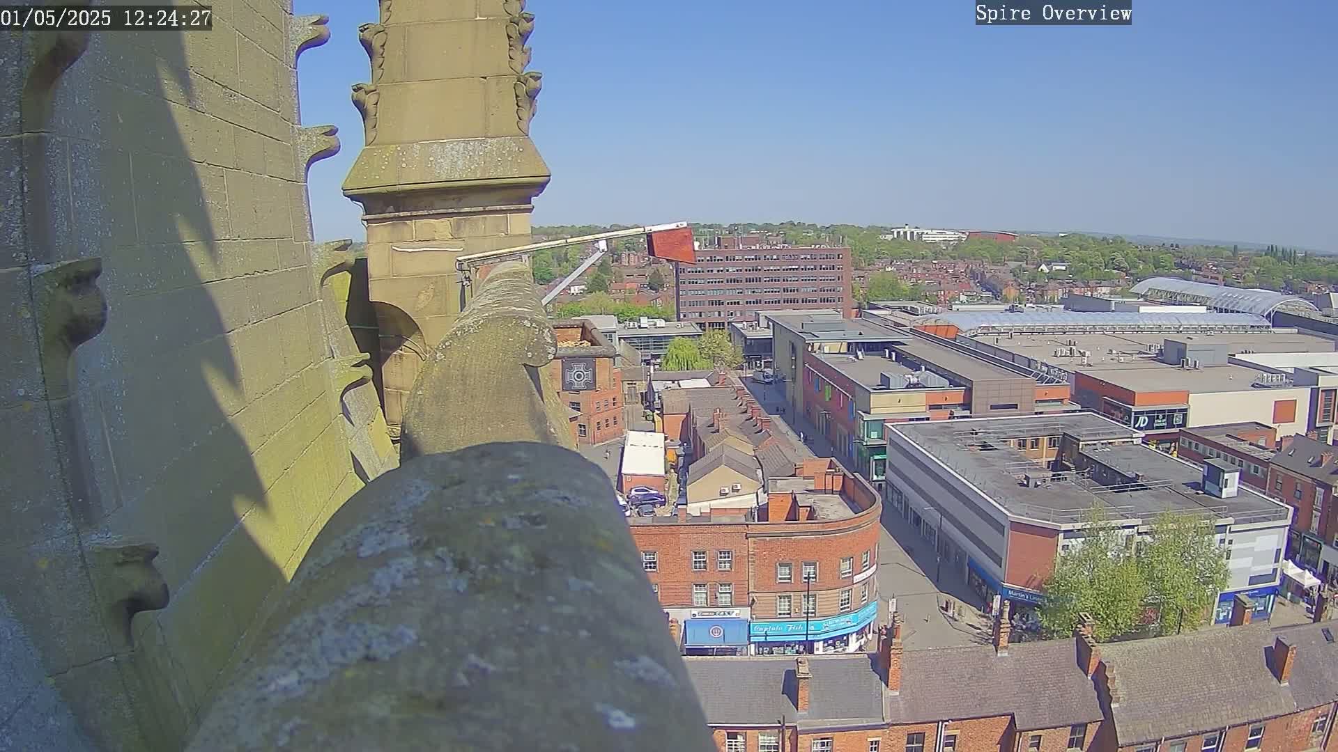 A high-angle view on a sunny day shows a stone structure, possibly a church spire, overlooking a town with a shopping center and many brick buildings.