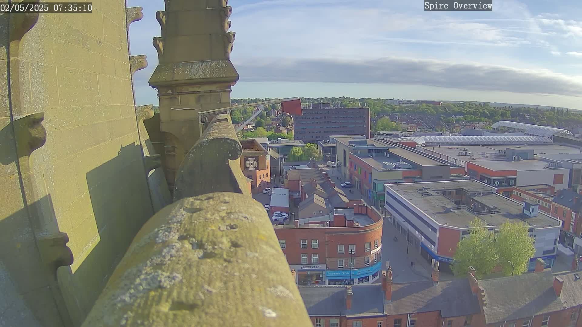 A high-angle view from a stone structure shows a partly sunny cityscape with a shopping center below, under a mostly clear sky.