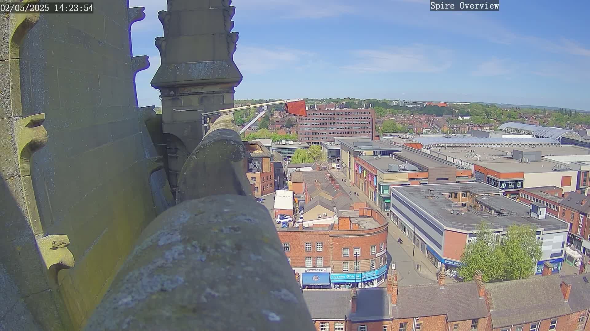 A high-angle view on a sunny day shows a city center with brick buildings and a large shopping mall, seen from atop a stone structure.