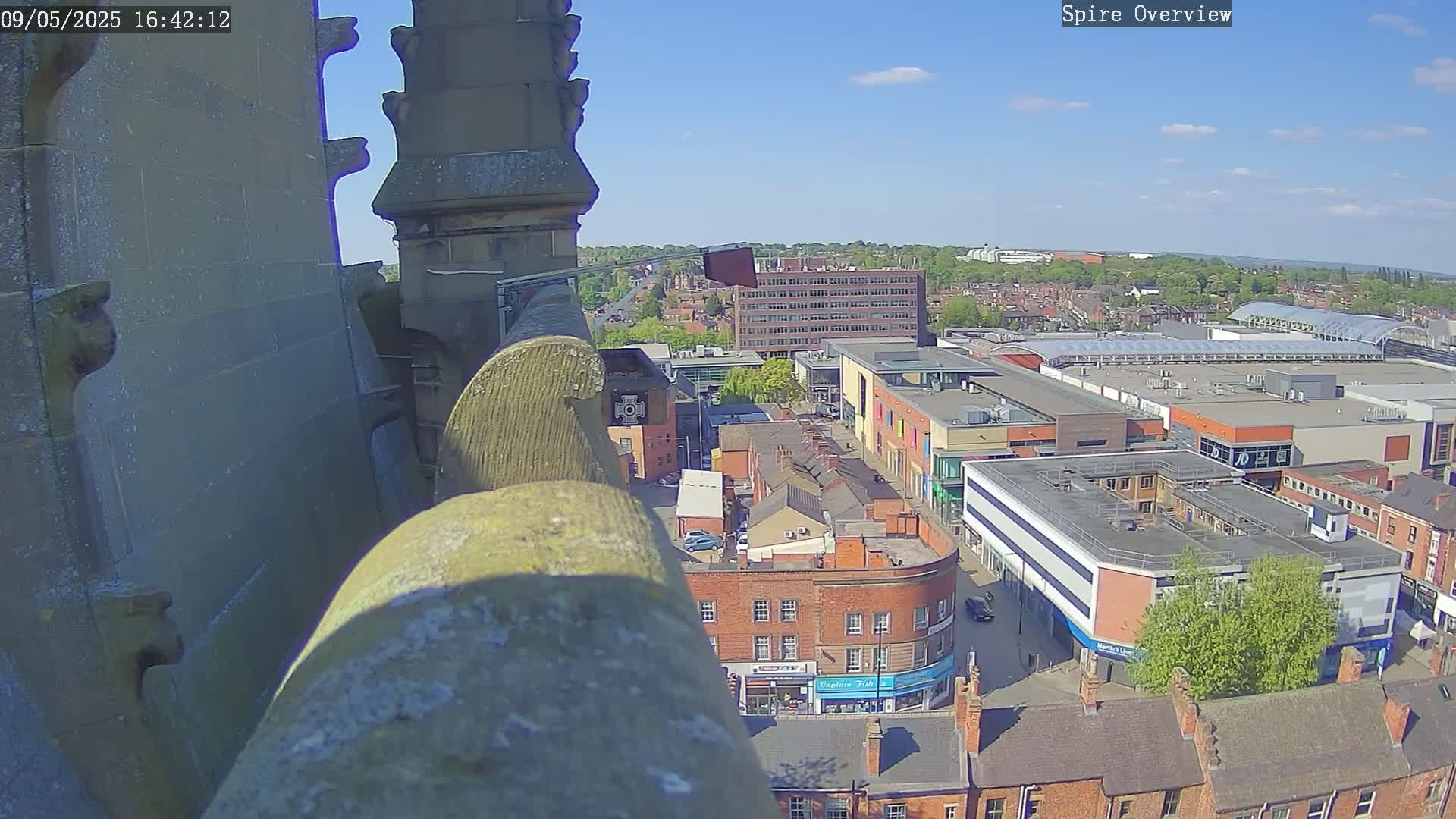 A high-angle, sunny view from a stone structure shows a town's cityscape with a shopping center and other buildings.