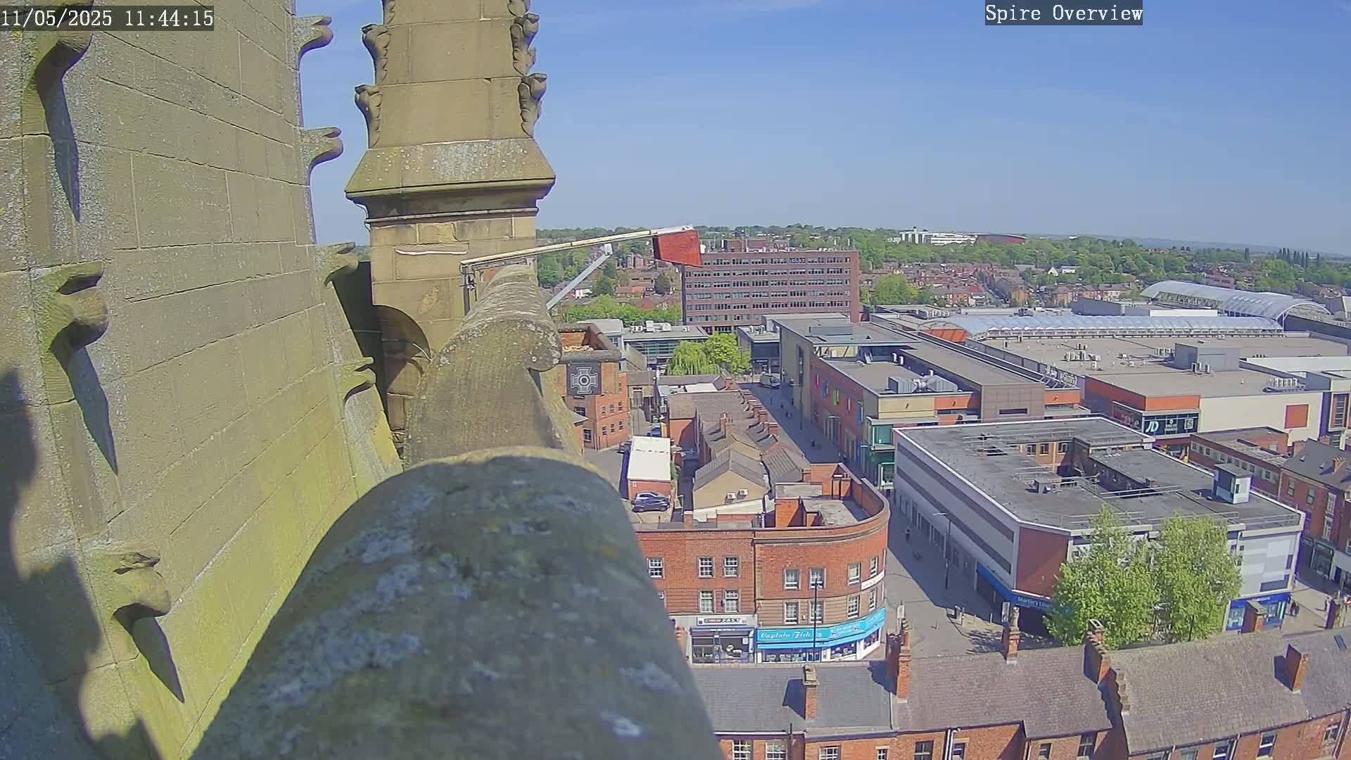 A high-angle view on a sunny day shows a city with brick buildings, a shopping mall, and a partially visible crane from a stone structure's rooftop.