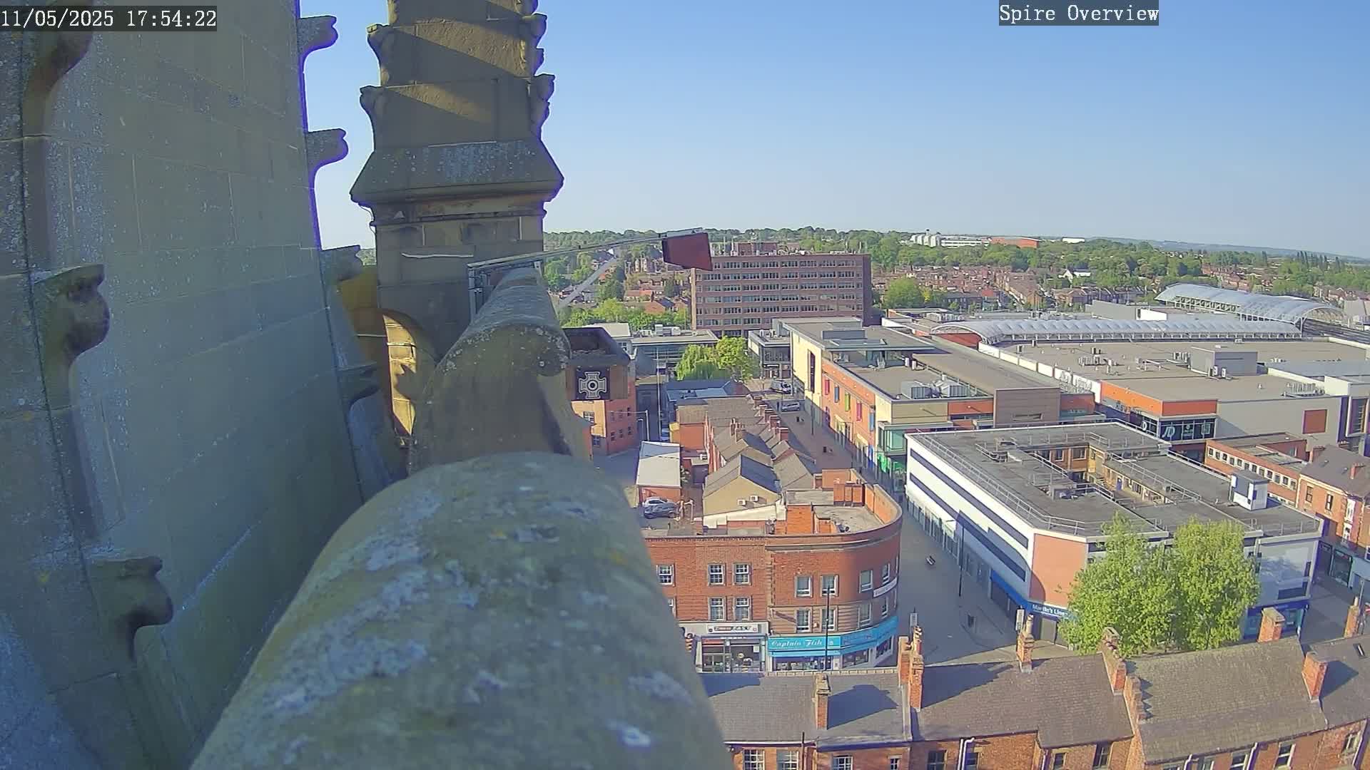 A high-angle view from a stone spire shows a town square with buildings and a shopping center under a clear, sunny sky.