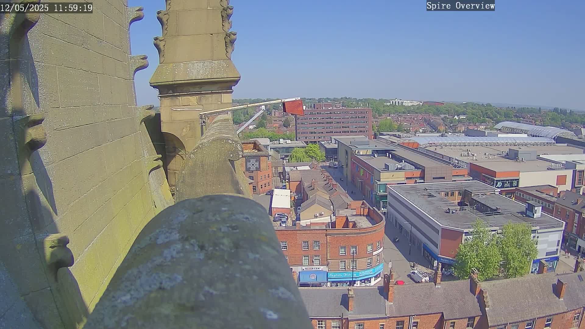 A high-angle view on a sunny day shows a cityscape with a shopping center, brick buildings, and a worker on a lift attached to a tall stone structure.