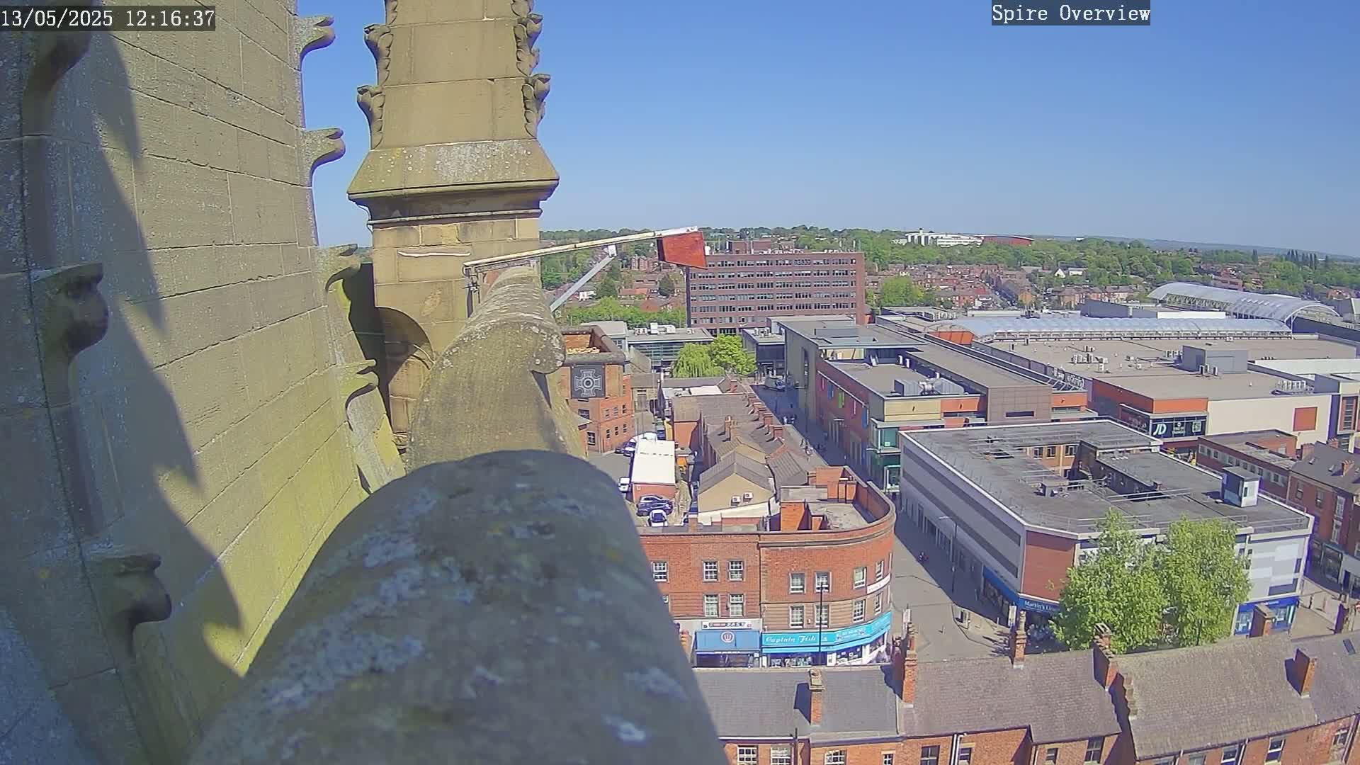 A high-angle view from a stone structure on a sunny day shows a city center with brick buildings, a large shopping mall, and a working lift or crane extending from the structure.