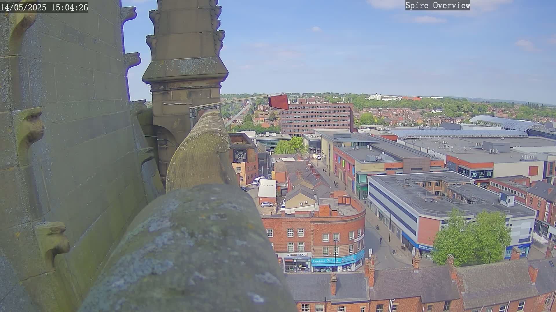 A high-angle, sunny view from a stone spire overlooking a town with a large shopping center and many brick buildings.