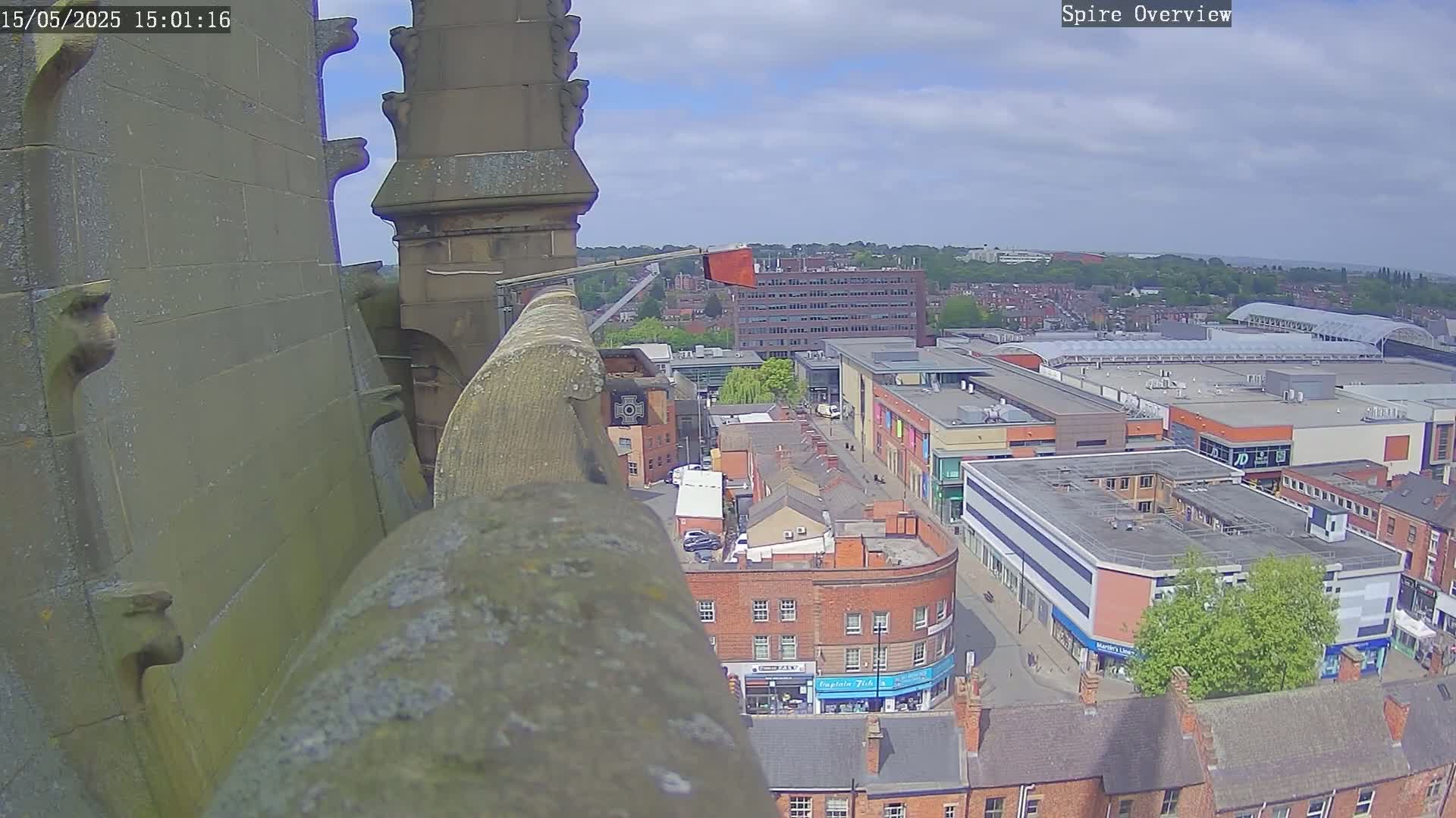 A high-angle view from a stone spire shows a partly sunny day over a city with brick buildings, a shopping center, and some green spaces.