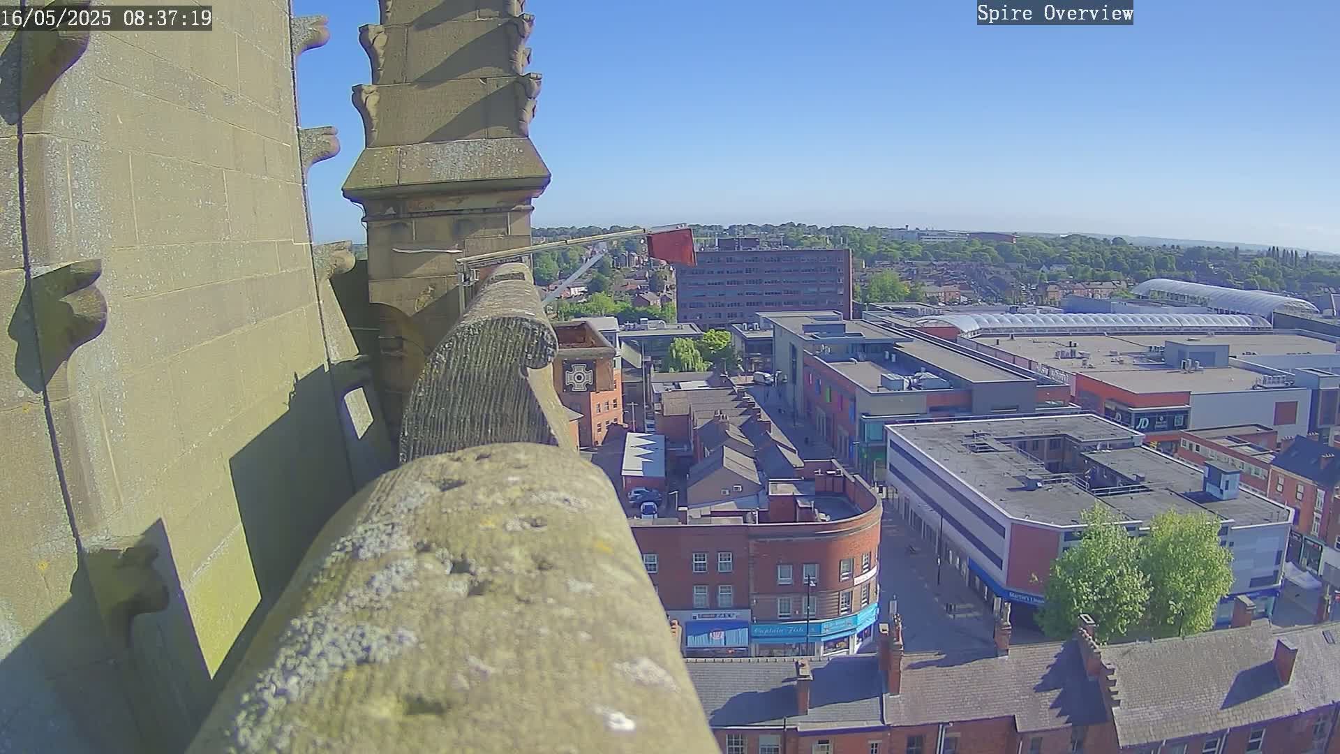 A high-angle, sunny view from atop a stone structure overlooks a town square with shops and a large shopping center.