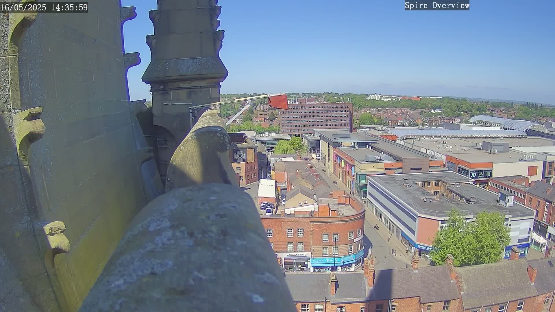 A high-angle view on a sunny day shows a city street scene with brick buildings and a large shopping center, seen from a stone structure's edge.