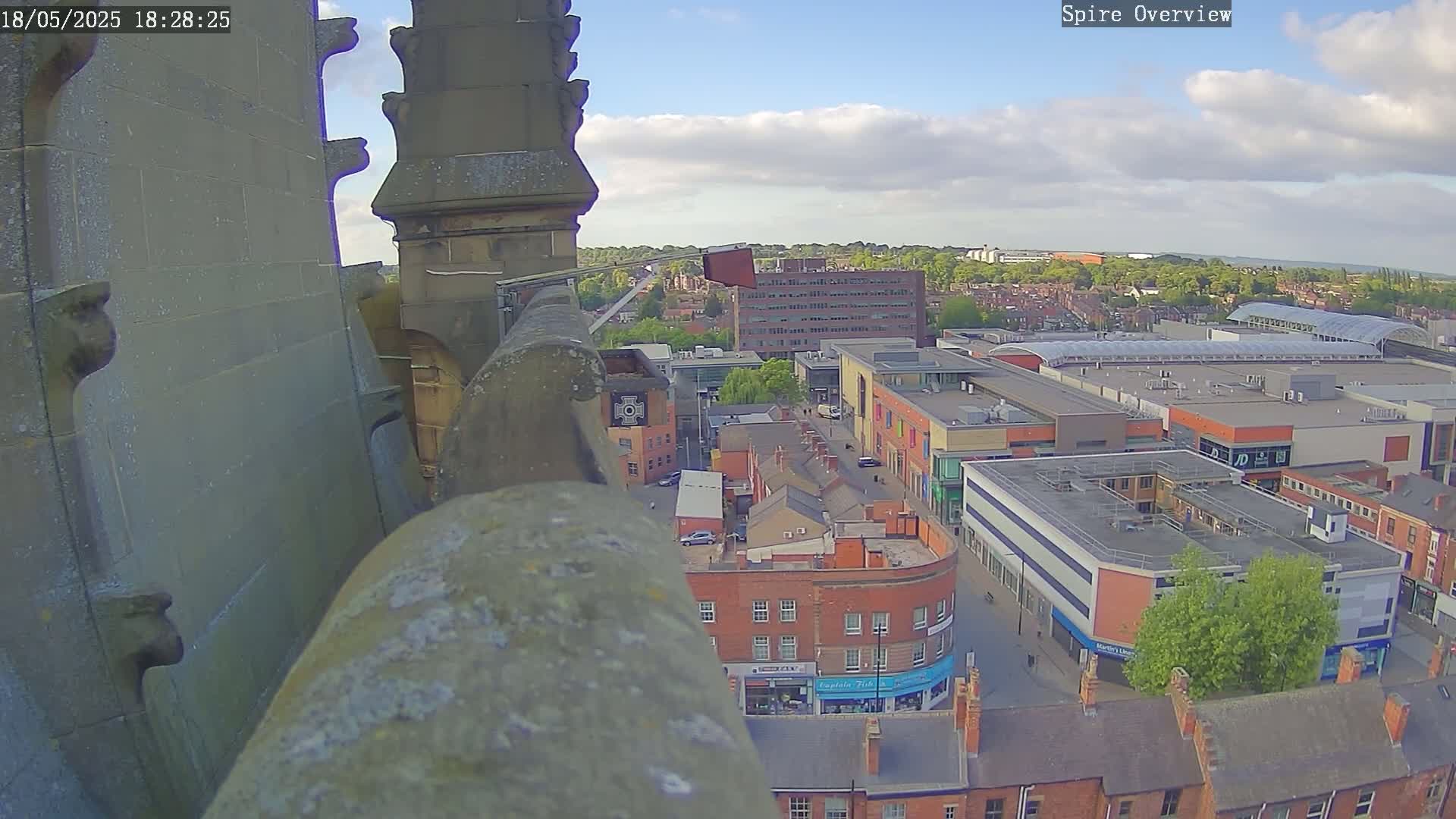 A high-angle view from a stone structure shows a city center with a shopping mall under partly cloudy skies.