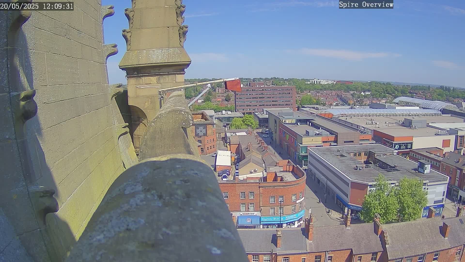 A high-angle, sunny view from a stone structure overlooking a city with brick buildings, a shopping center, and some trees.