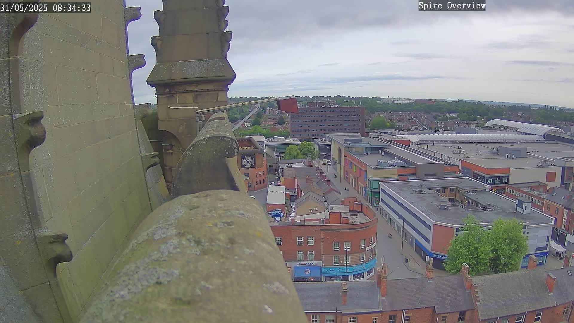 A high-angle view from a stone structure shows an overcast day over a town's cityscape, including a large shopping center and brick buildings.