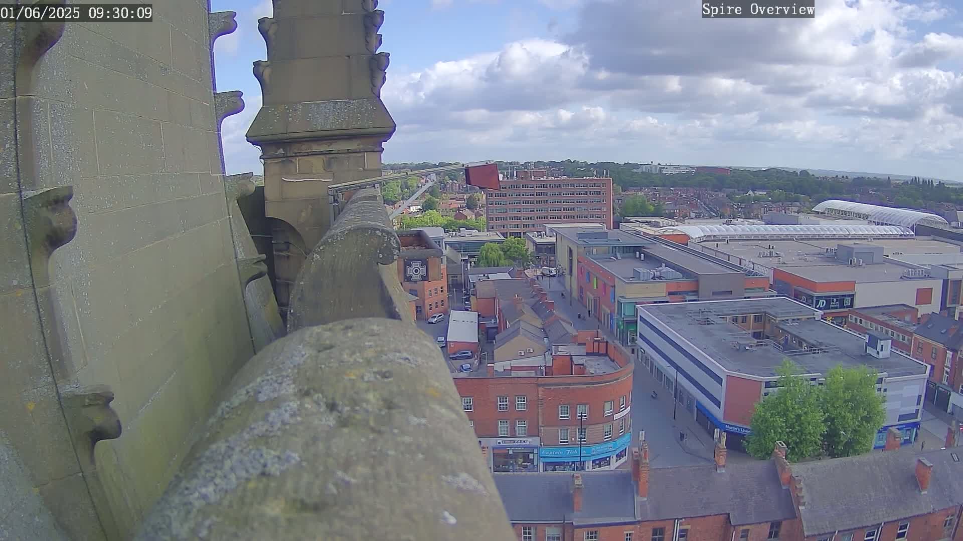 A high-angle view from a stone structure shows a partly cloudy day over a city with buildings and streets visible below.