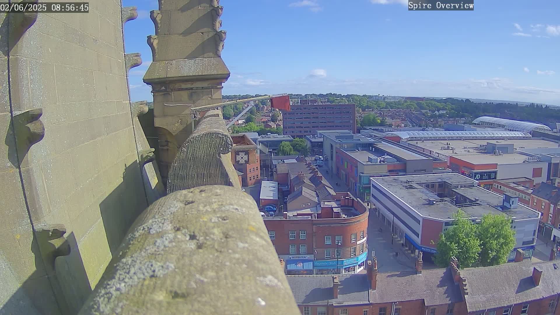 A high-angle, sunny view from a stone structure shows a town with shops and buildings below, under a mostly clear blue sky.