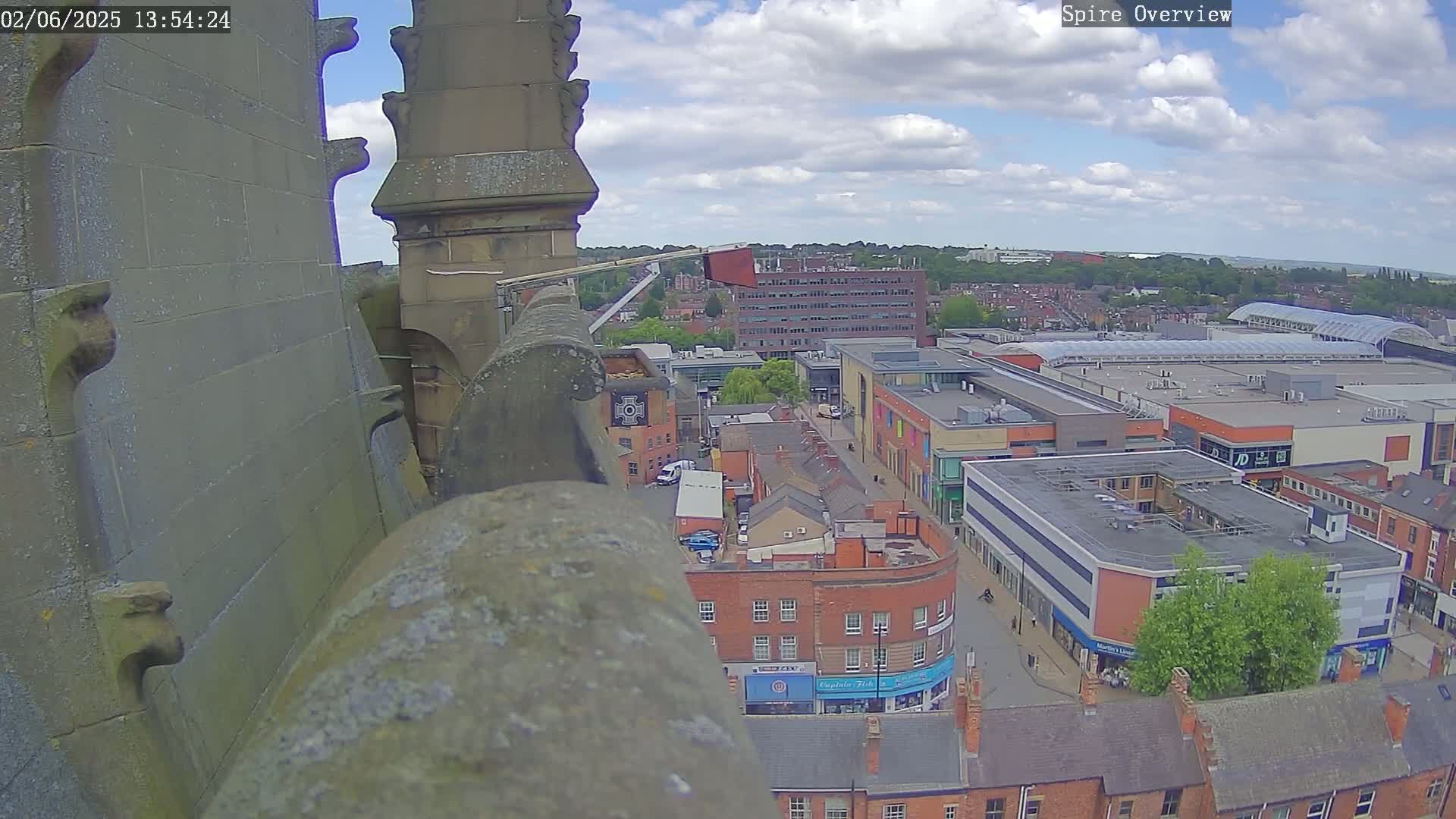 A high-angle view from a stone structure shows a partly cloudy day over a town with a shopping center and many brick buildings.