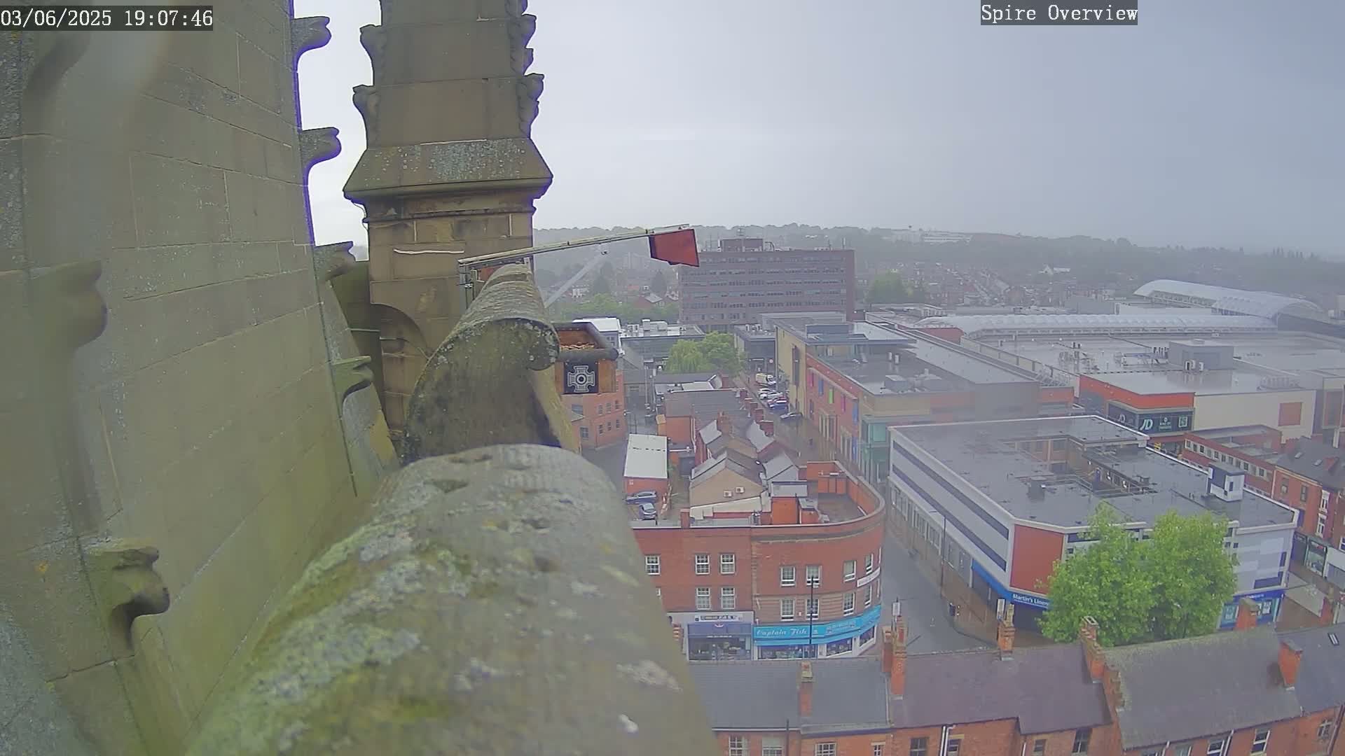 A high-angle view from a stone structure shows a town under an overcast sky, with rain falling on buildings and streets.
