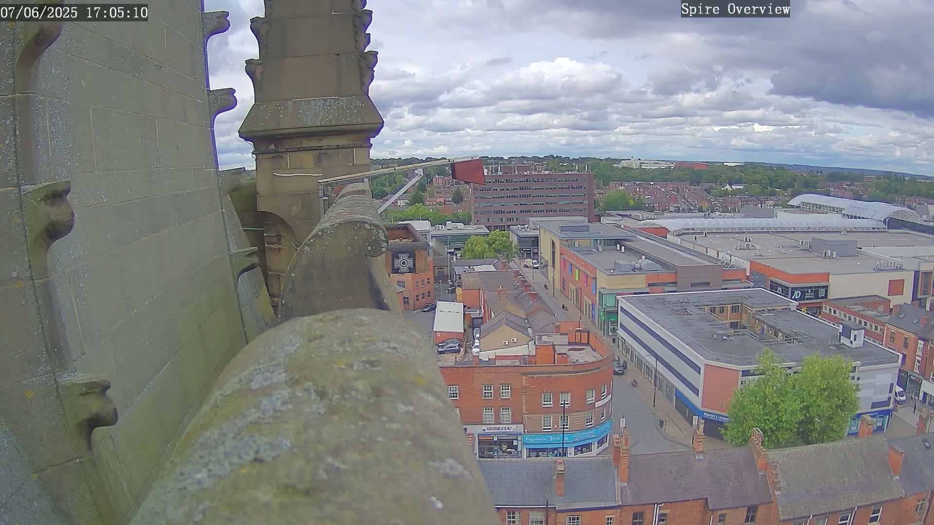 A high-angle view from a stone structure shows a town under partly cloudy skies, with a shopping center prominent in the distance.