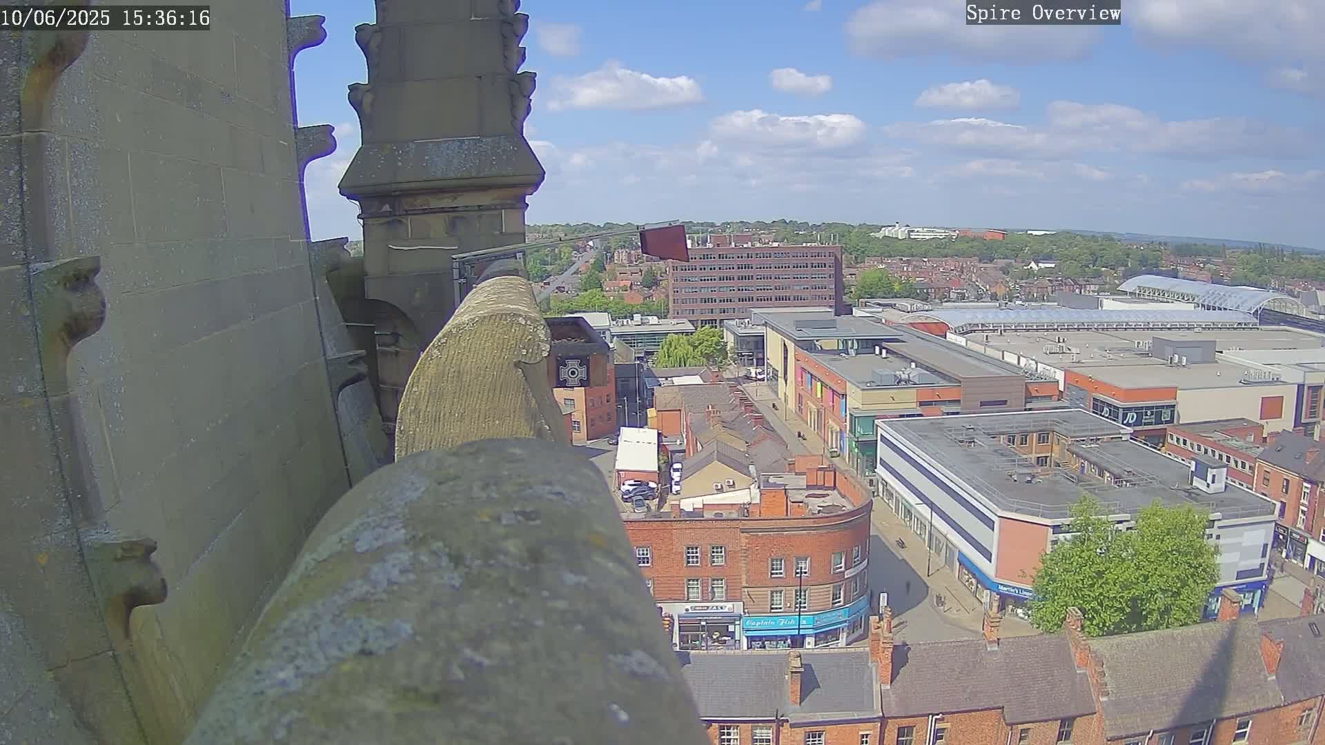 A high-angle view from a stone structure on a partly cloudy day shows a town square with buildings and a shopping center below.
