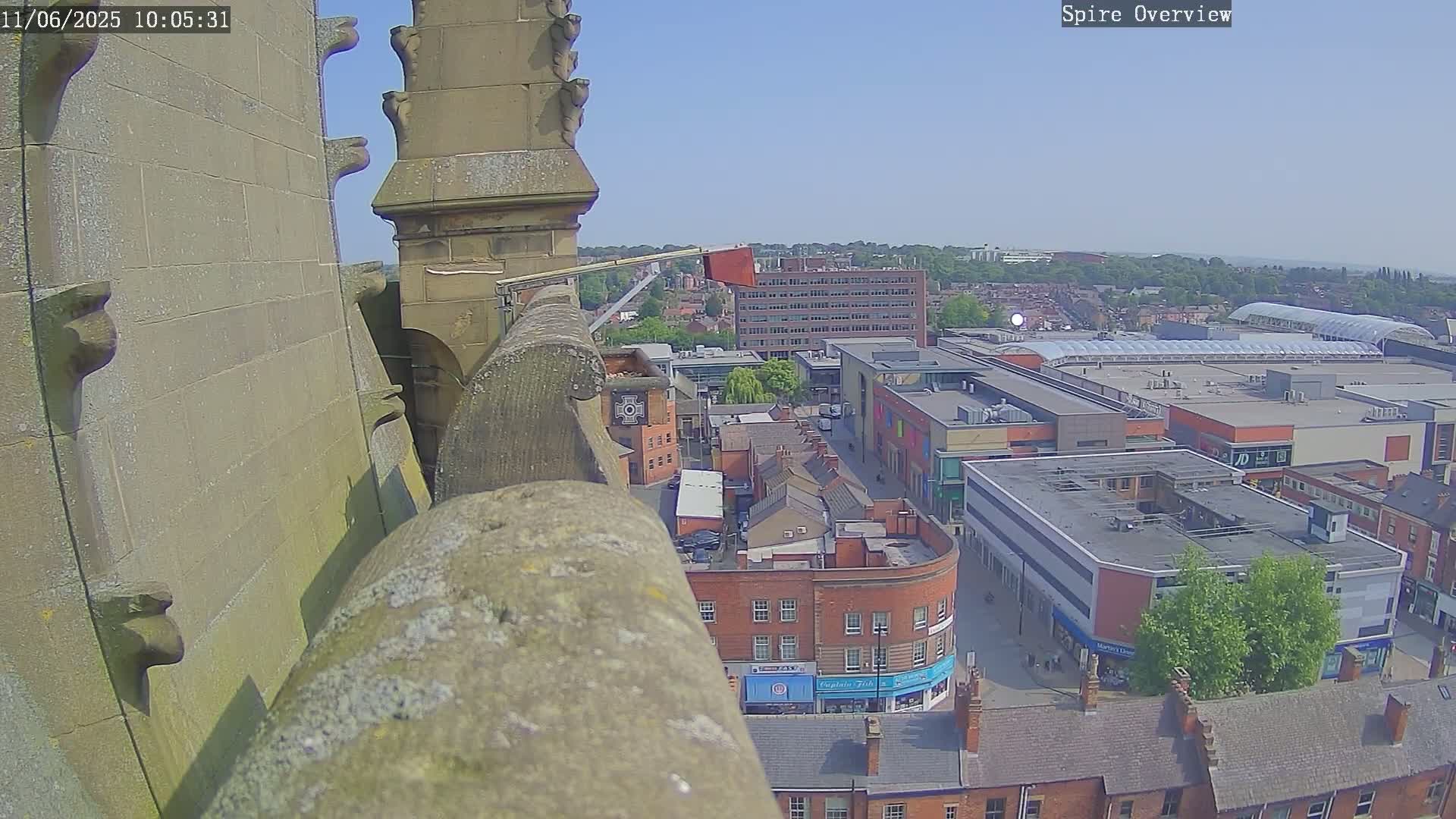 A high-angle, outdoor view on a sunny day shows a stone structure overlooking a town square with brick buildings and a large shopping mall.