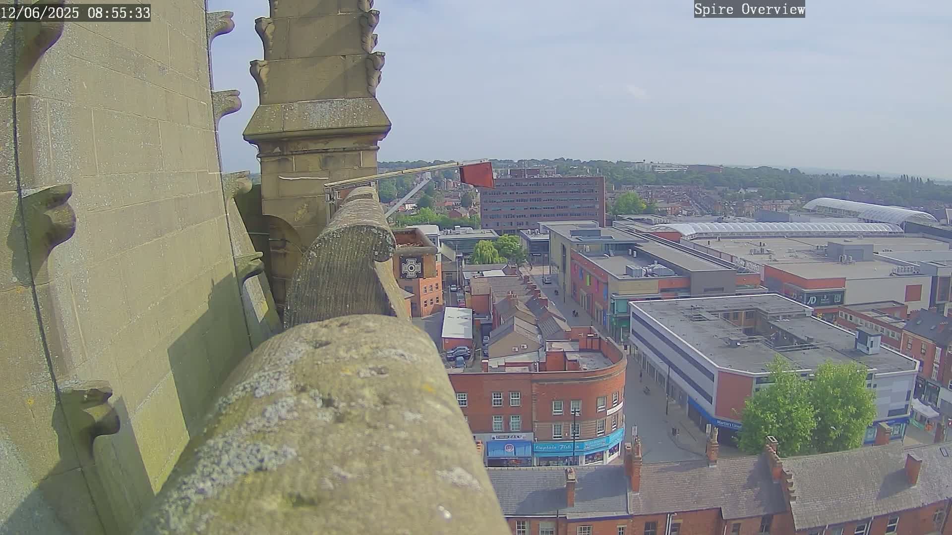 A high-angle view on a sunny day shows a stone spire overlooking a town with shops and buildings below.