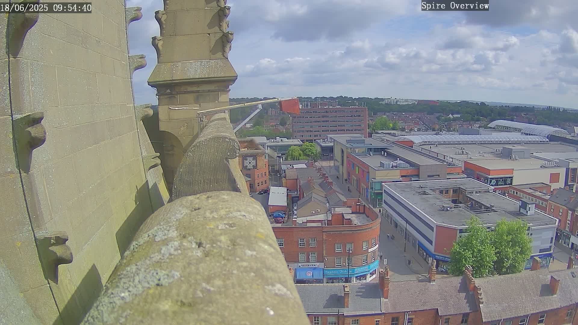 A high-angle view from a stone structure shows a partly cloudy day over a town with a shopping center, featuring a mix of brick buildings, roads, and green spaces.