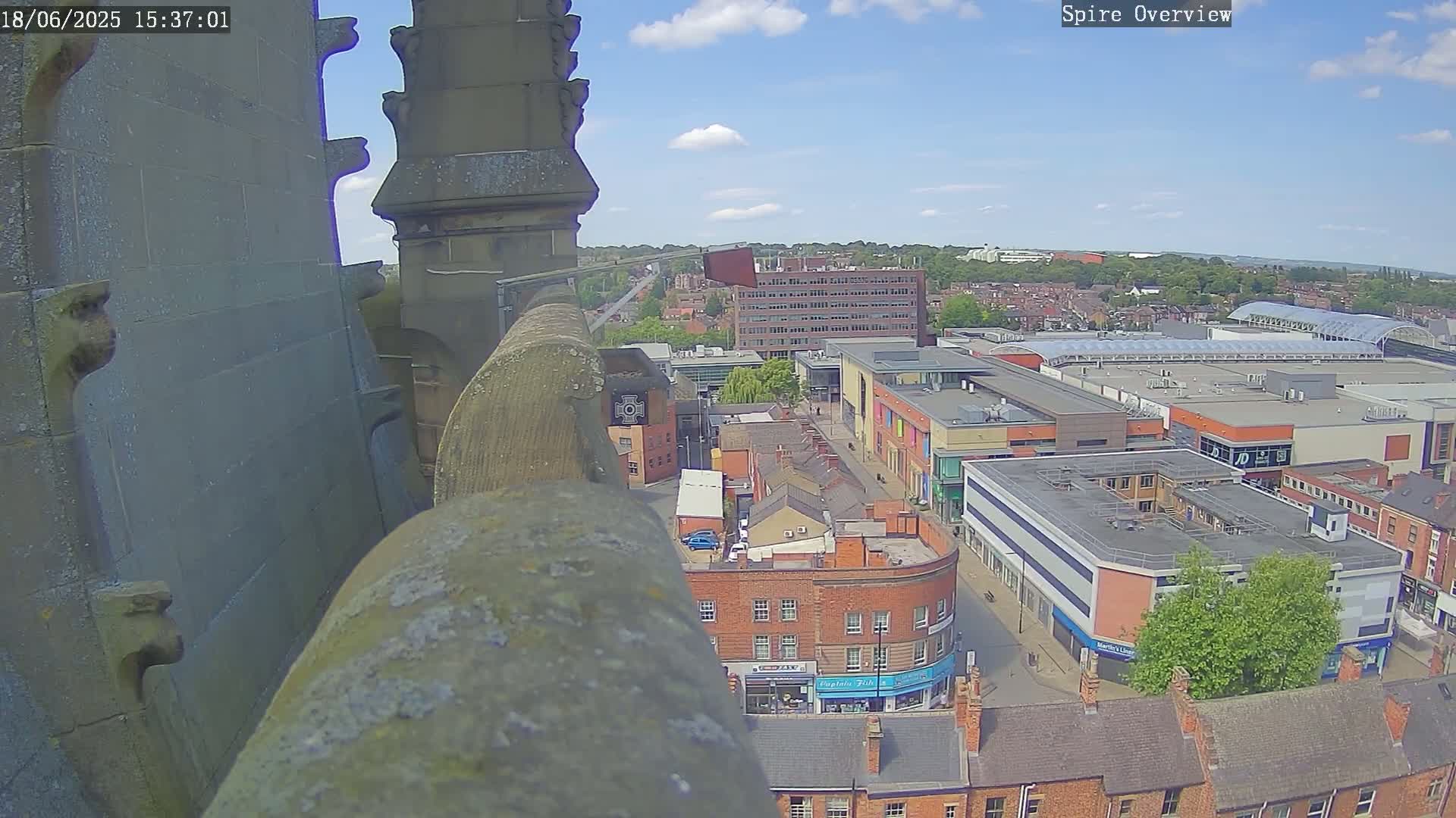 From a high vantage point on a sunny day, a view overlooks a town square with brick buildings, shops, and a large modern shopping center, partially obscured by a stone structure in the foreground.