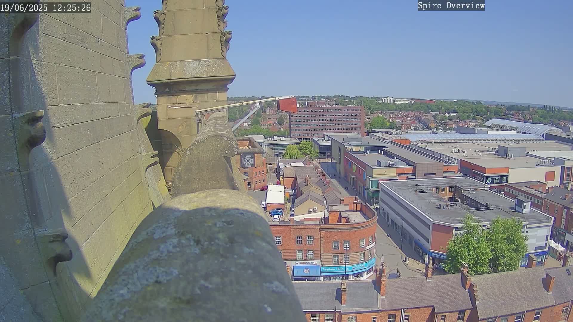 A high-angle view on a sunny day shows a stone spire overlooking a town with brick buildings, a shopping center, and tree-lined streets.