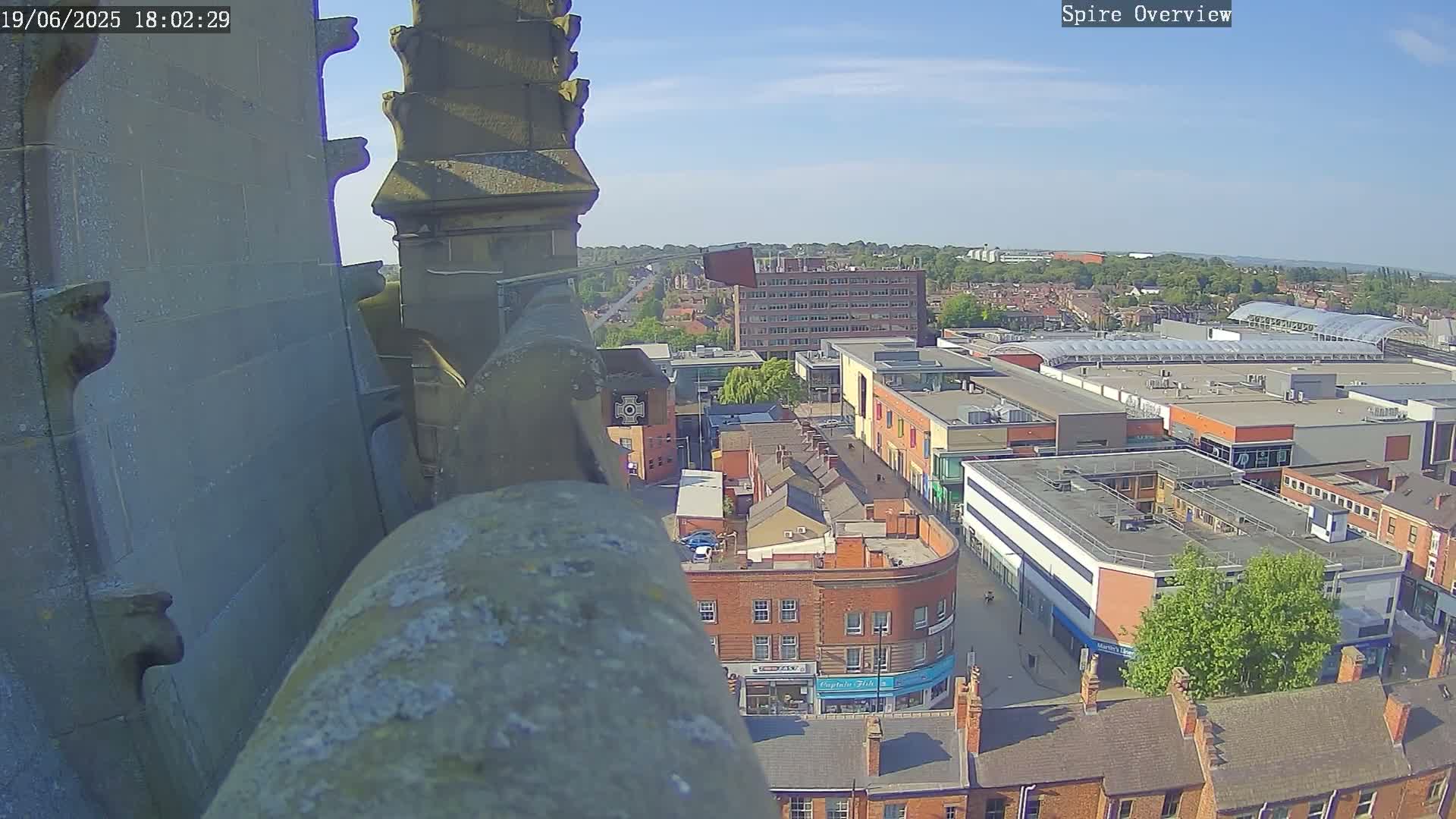 A high-angle, outdoor view on a sunny day shows a town center with brick buildings and a shopping mall, seen from atop a stone structure.
