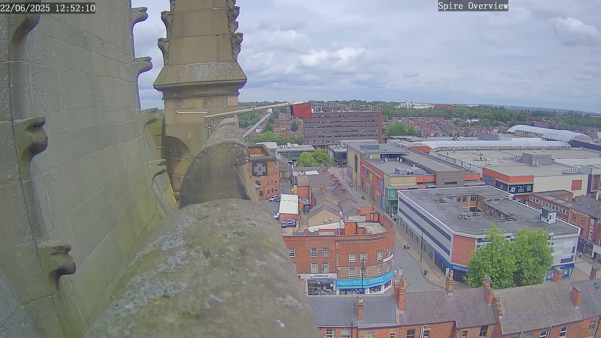 A high-angle view from a stone structure shows a partly cloudy sky over a town with buildings and streets below on a partly sunny day.