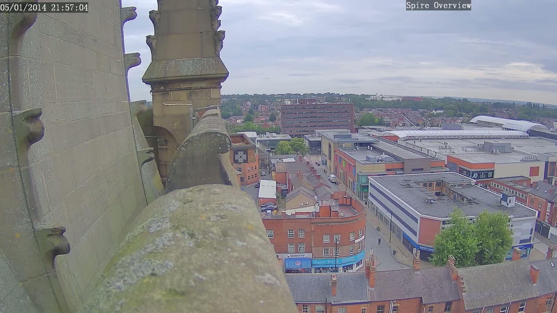 A high-angle view from a stone structure shows an overcast day over a city with brick buildings and a large shopping center.