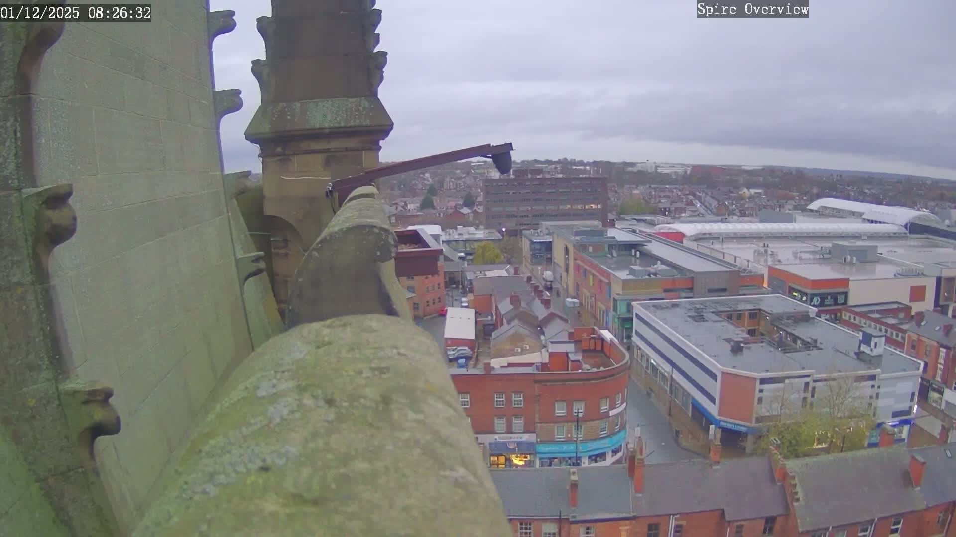 An overcast and gloomy high-angle view from a stone spire looks down upon a sprawling city with a mix of commercial and residential buildings, featuring a large shopping complex, streets, and scattered trees.