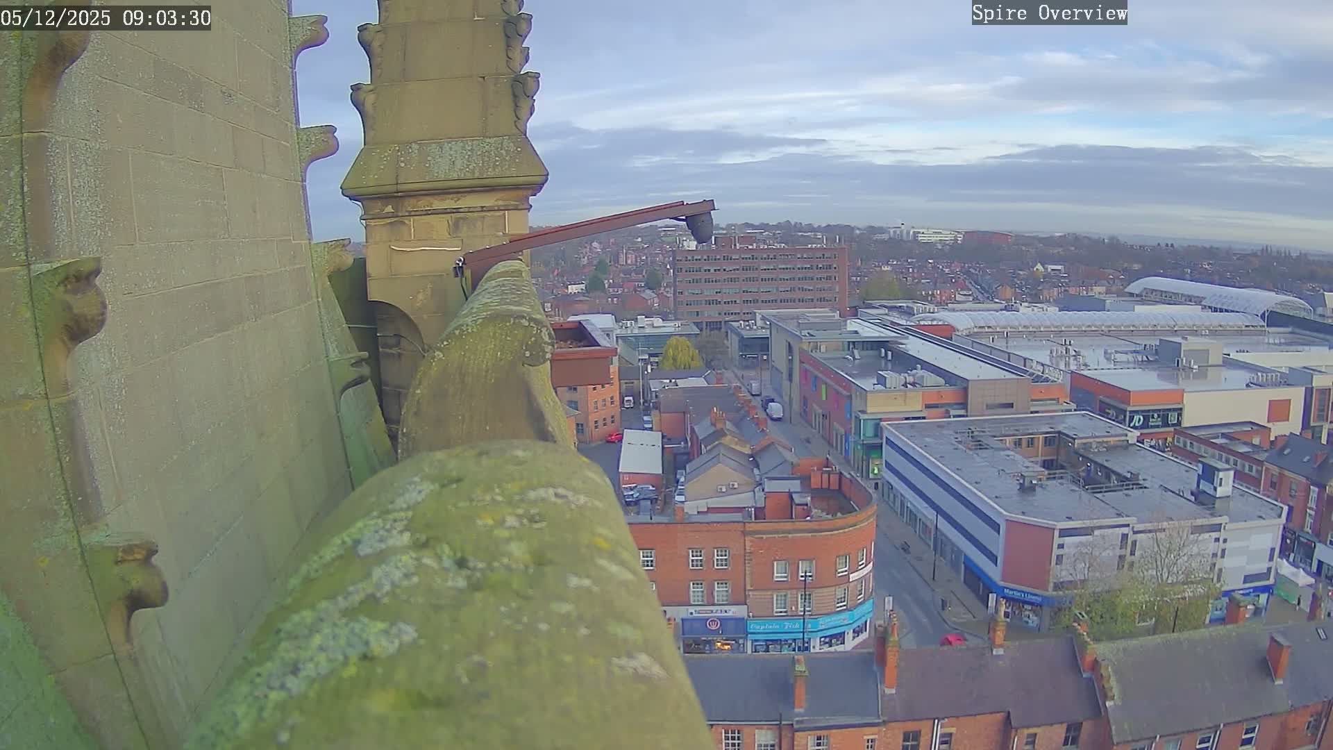 The image captures an overcast day overlooking an urban landscape from a moss-covered stone architectural structure, revealing a dense mix of traditional brick buildings, modern commercial complexes, and a large shopping center under a cloudy sky.