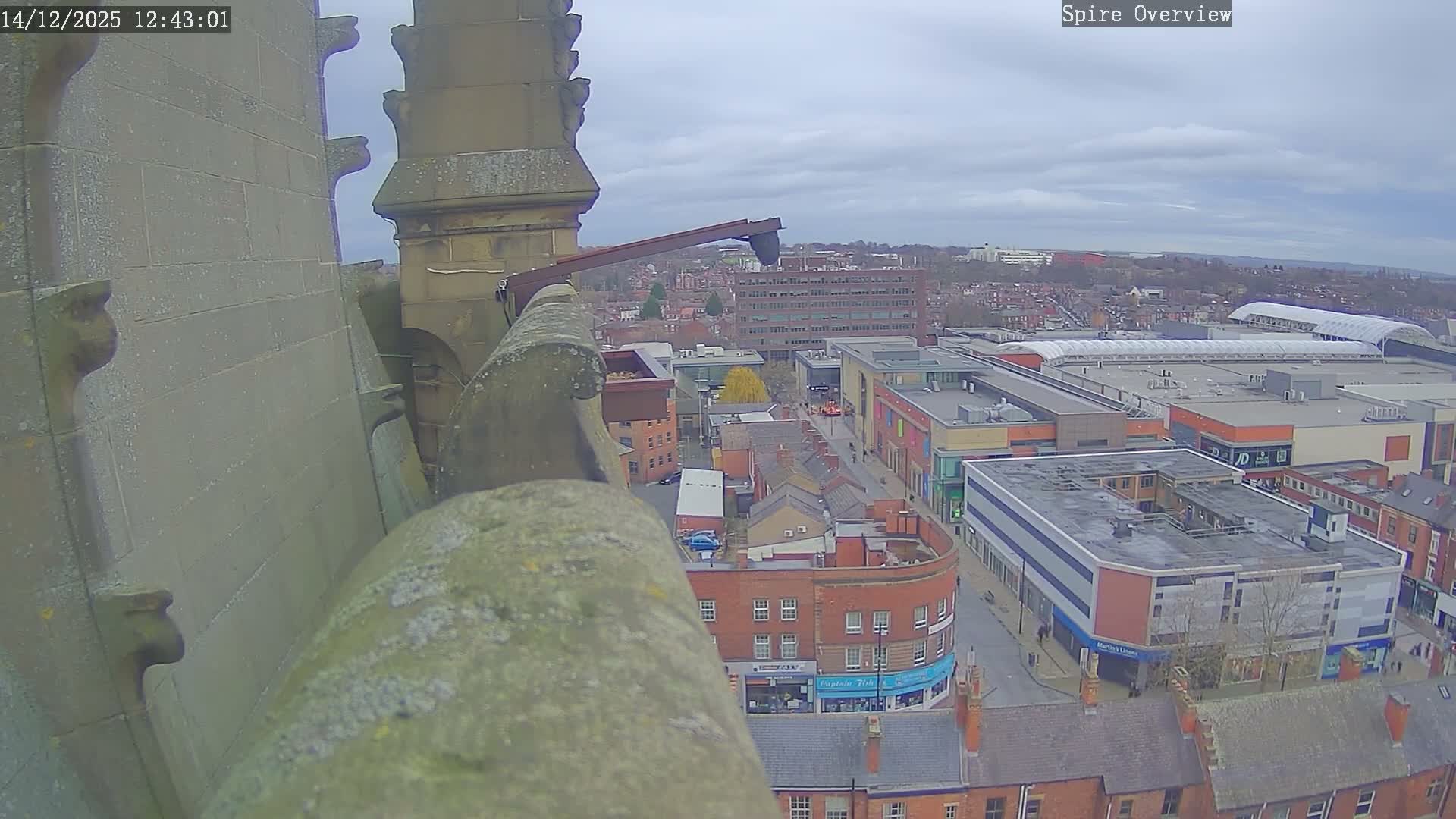 The image captures an overcast day overlooking an urban landscape from a moss-covered stone architectural structure, revealing a dense mix of traditional brick buildings, modern commercial complexes, and a large shopping center under a cloudy sky.
